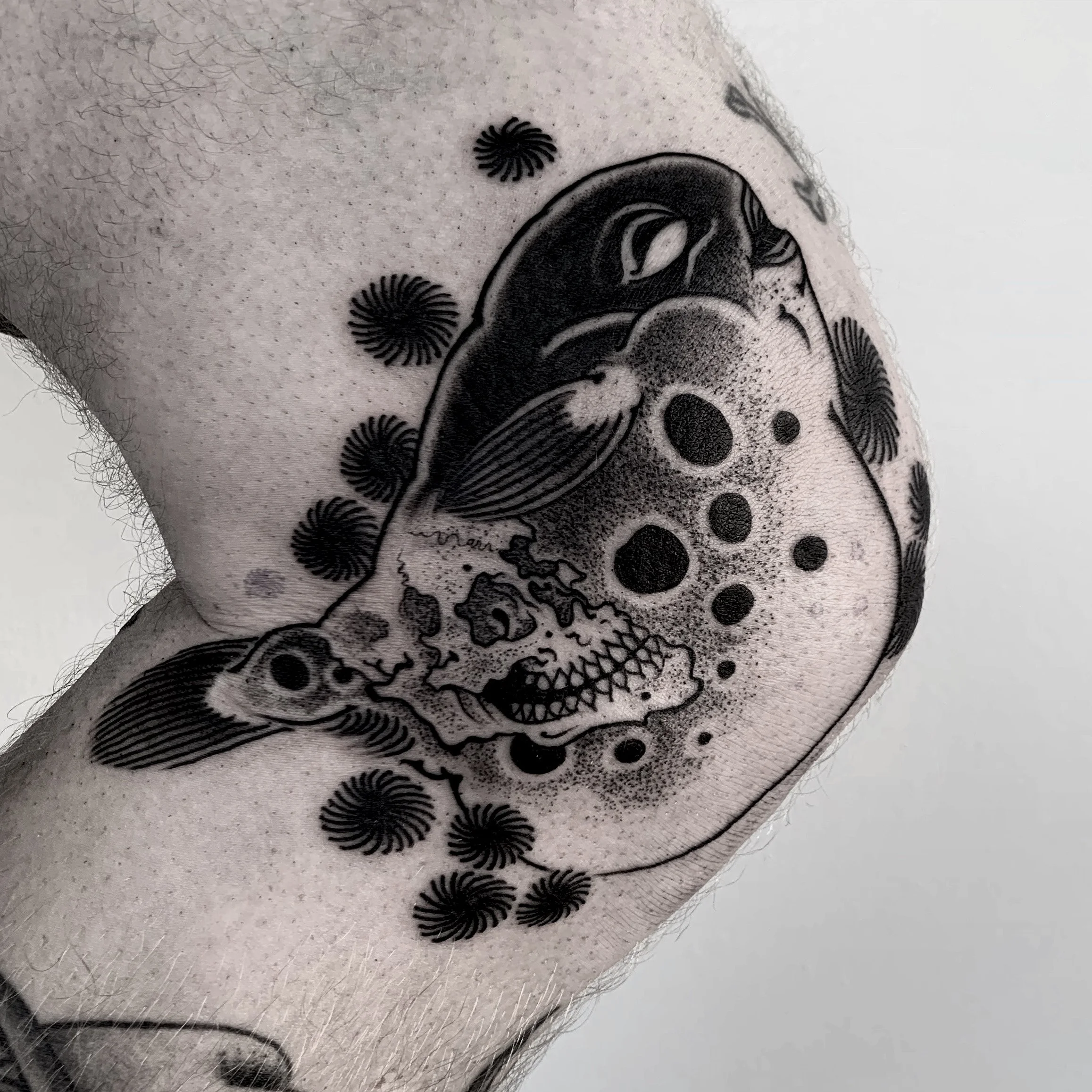 Black and gray tattoo of a skull, a fish, and a hockey mask on a person's arm, with some circular patterns.