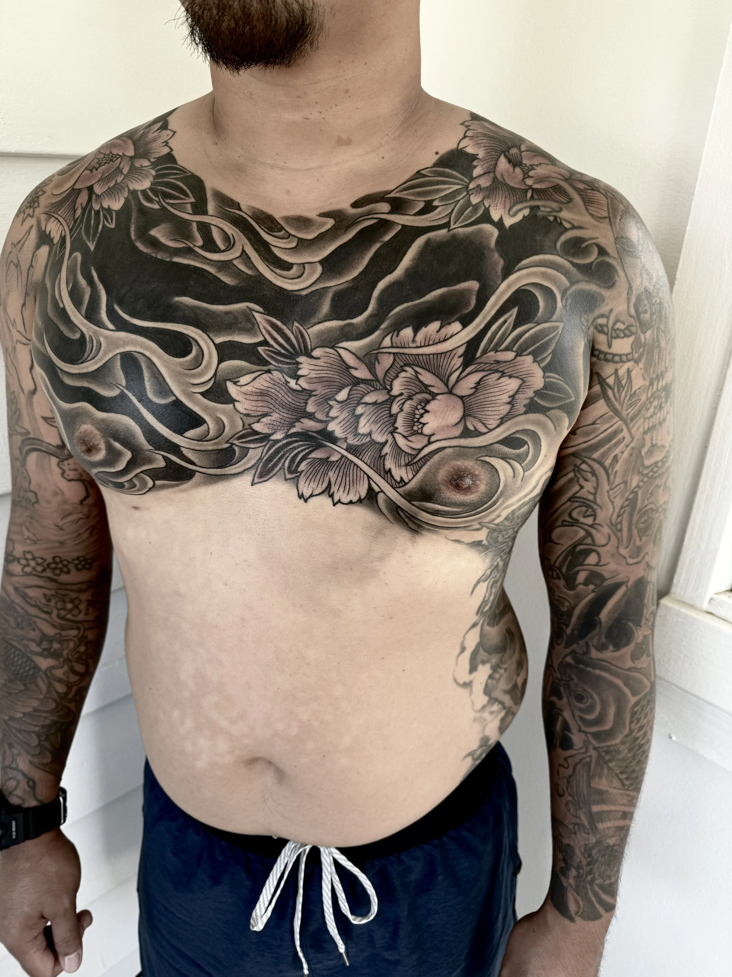 A shirtless man with extensive black and grey tattoos covering his chest, arms, and stomach, standing indoors.