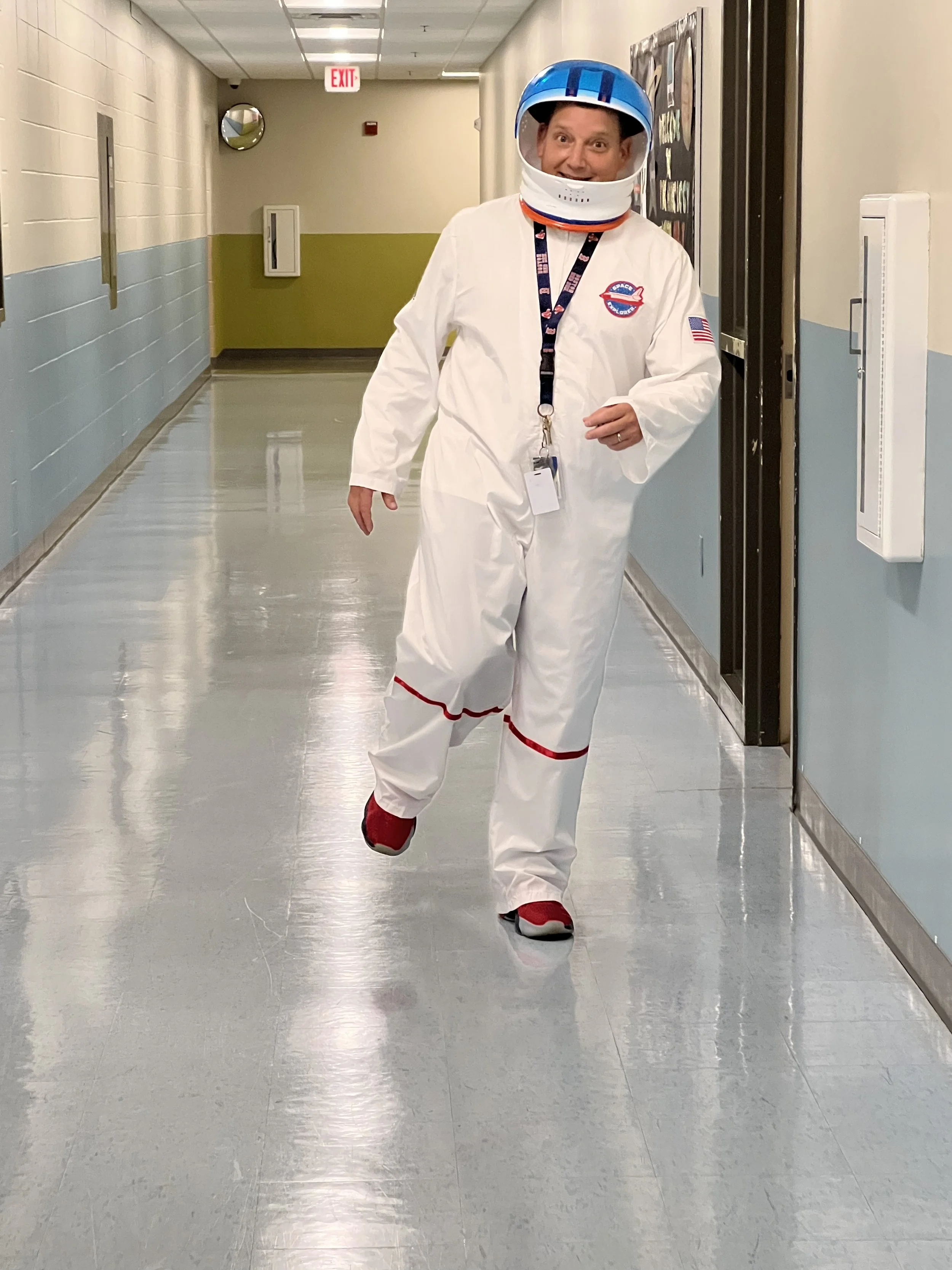 A person dressed in a white NASA astronaut suit with a blue helmet, walking down a hospital corridor, smiling, with an exit sign and wall decor in the background.