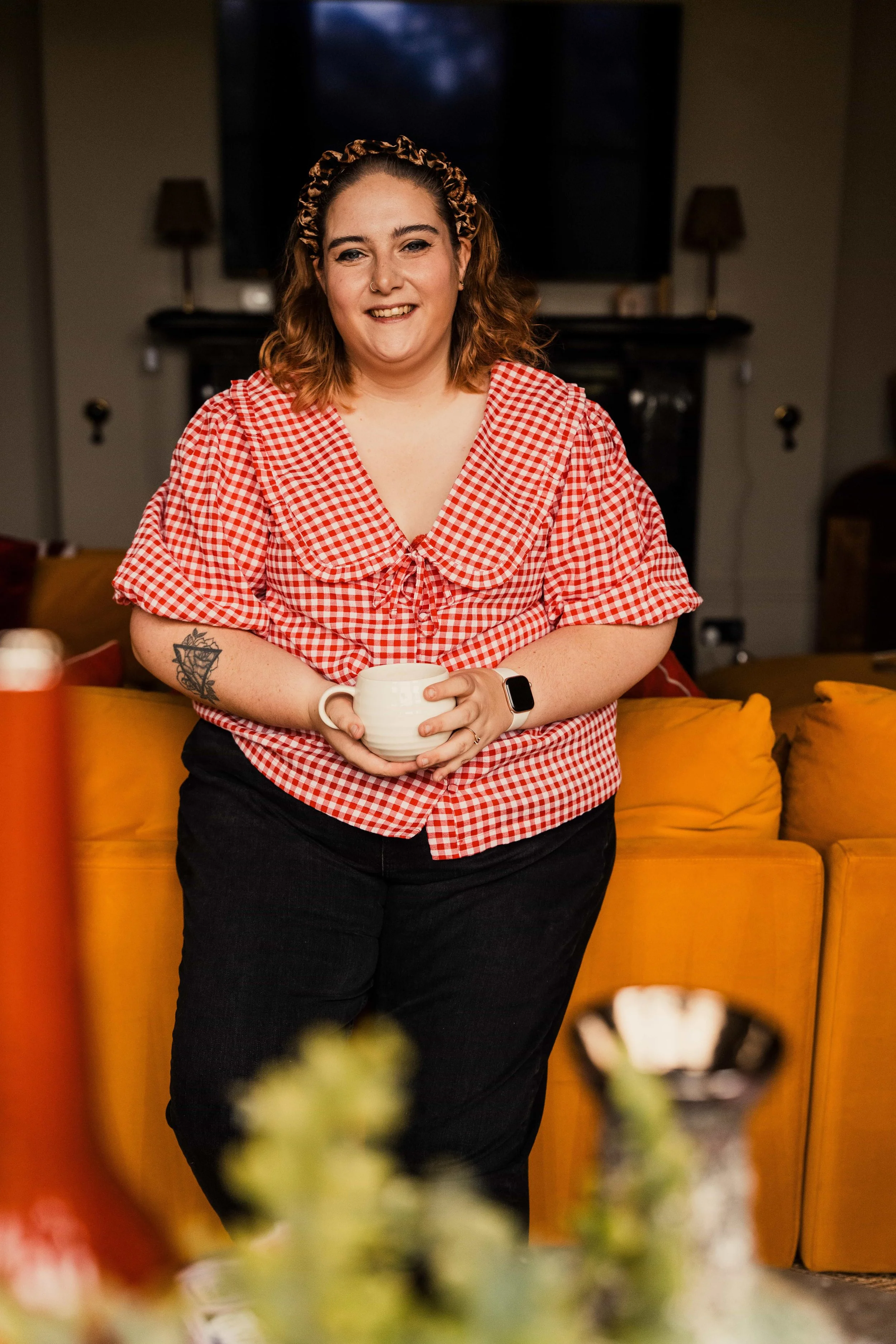 woman smiling wearing headband and red checked top holding a cup leaning against an orange sofa