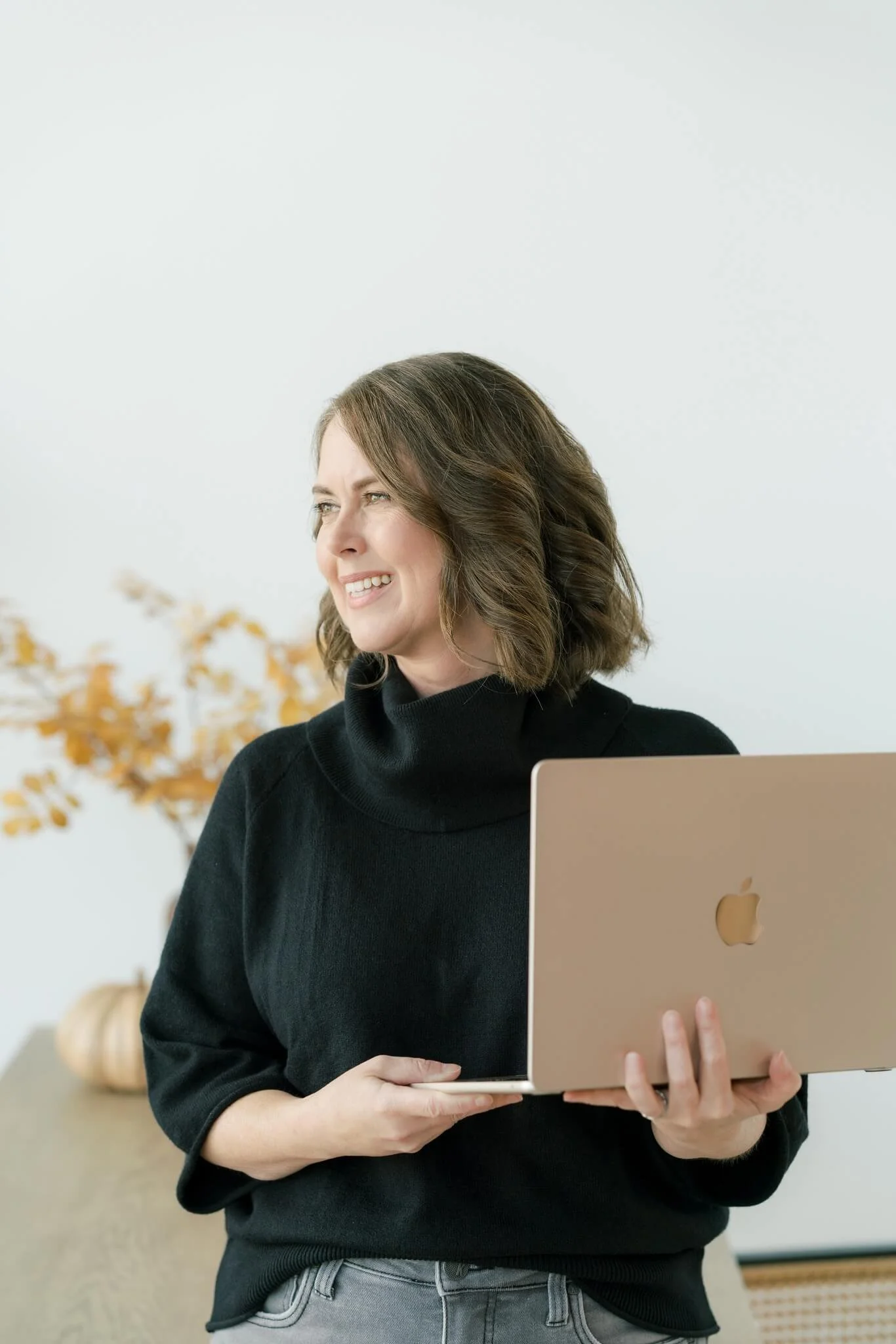 Woman with shoulder-length brown hair holding a gold MacBook, smiling and looking to the side indoors with a white wall and autumnal decorations in the background.