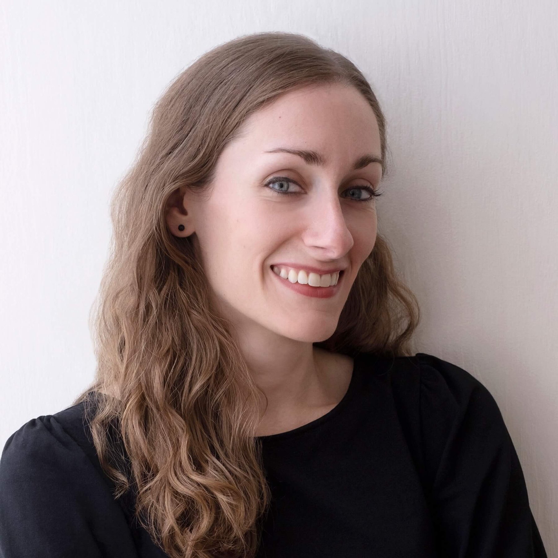 A woman with wavy brown hair, wearing a black top, smiling and looking at the camera, standing against a white wall.