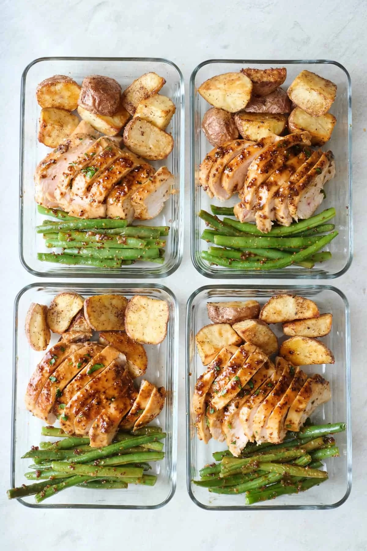 Four glass meal prep containers with grilled chicken breast topped with sauce, roasted potatoes, and green beans.