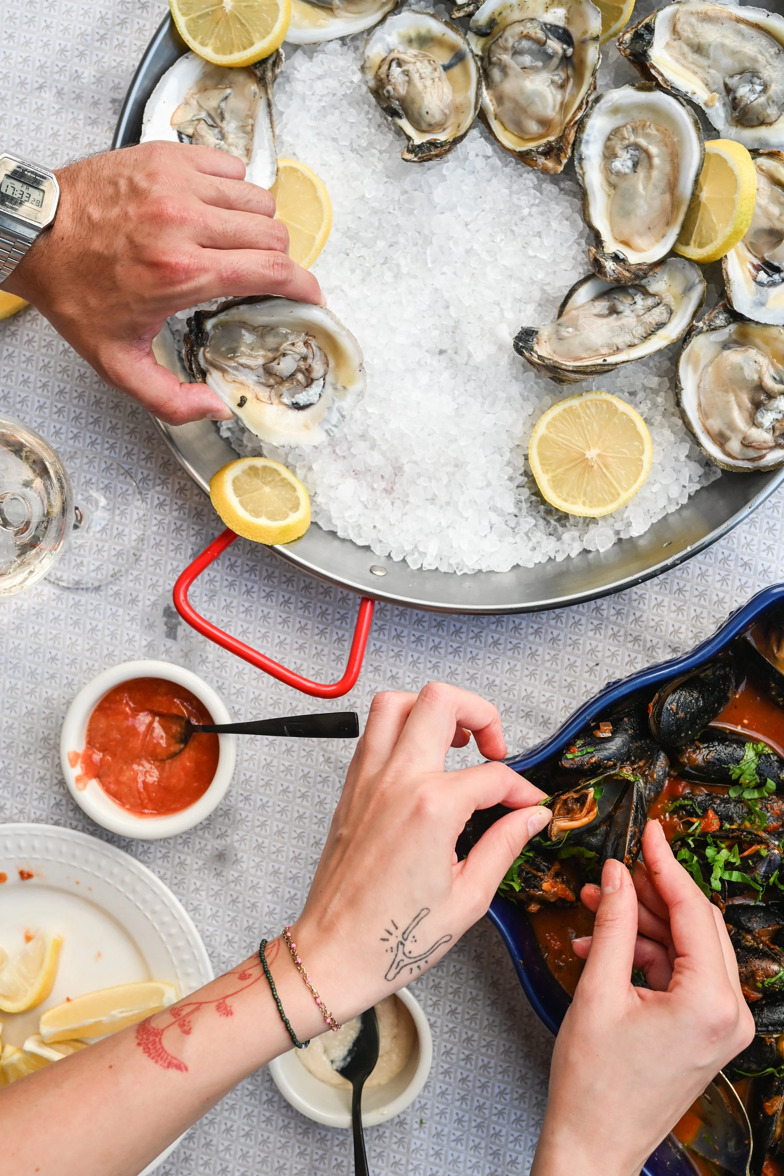 A seafood meal featuring fresh oysters on a bed of ice with lemon slices, a bowl of cocktail sauce, and a dish of mussels in a tomato-based sauce, with two people serving themselves.