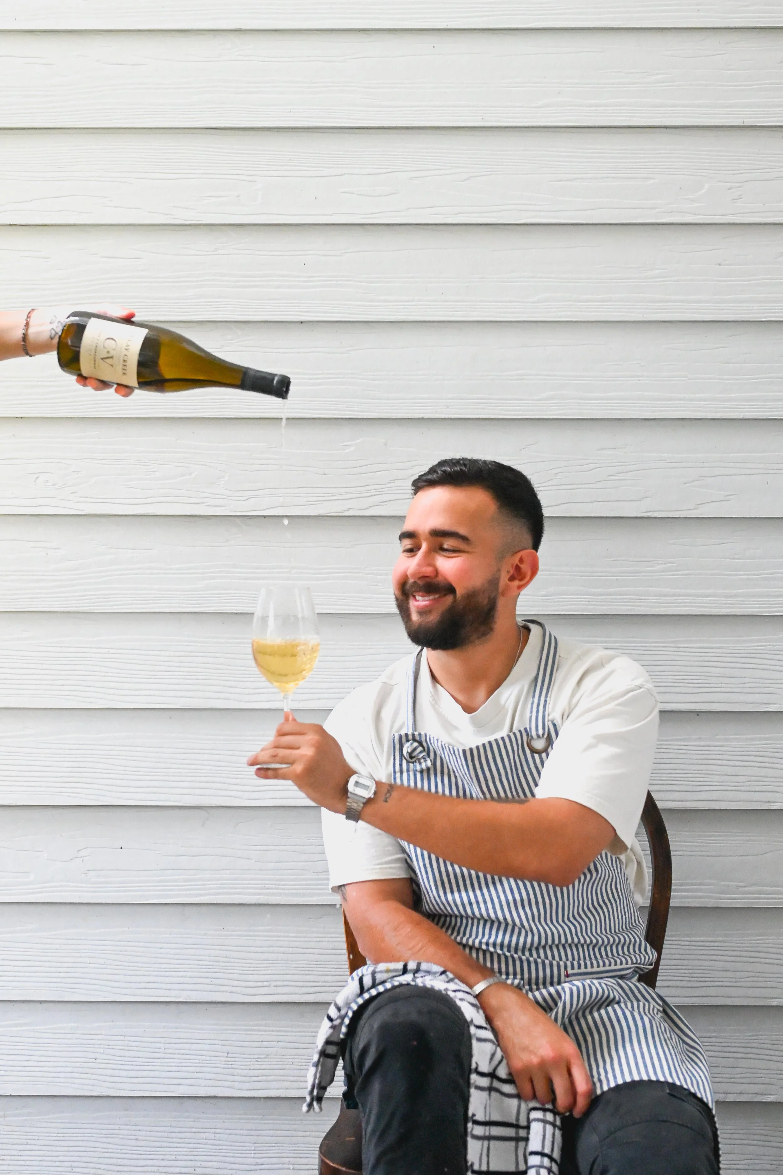 A man with a beard and short dark hair sitting on a wooden chair, holding a wine glass filled with white wine, while someone pours champagne from a bottle above him. The background is a white wooden wall, and the man is wearing a white shirt with an apron.