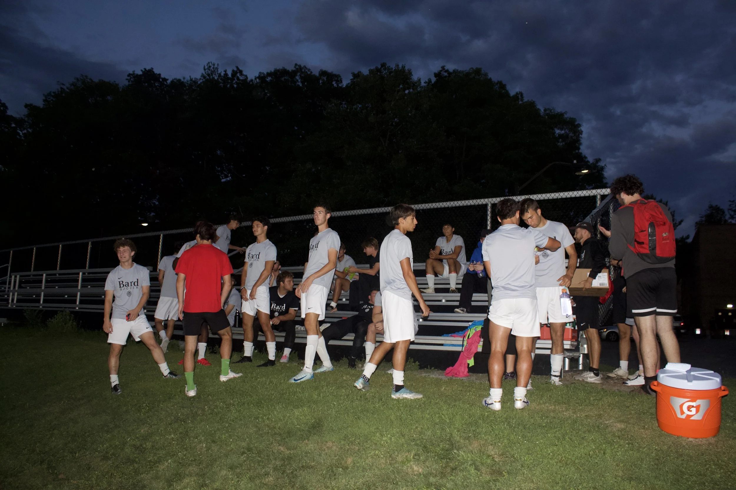 A group of young men dressed in white soccer uniforms with 'Bard Soccer' printed on their shirts, gathered on a grassy field near bleachers during dusk. Some are standing, others seated, with backpacks and water bottles nearby. Dark trees and a cloud