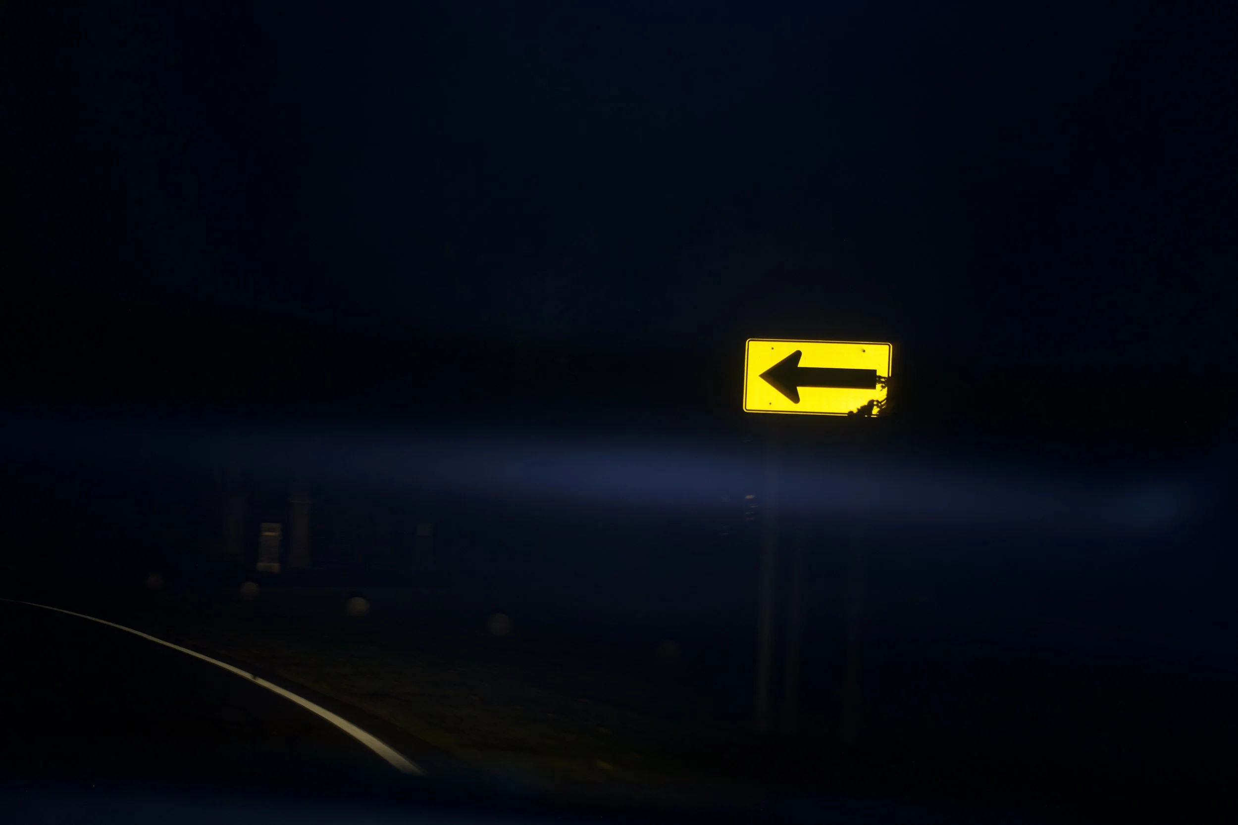 A yellow road sign with a black left arrow, illuminated at night against a dark sky.