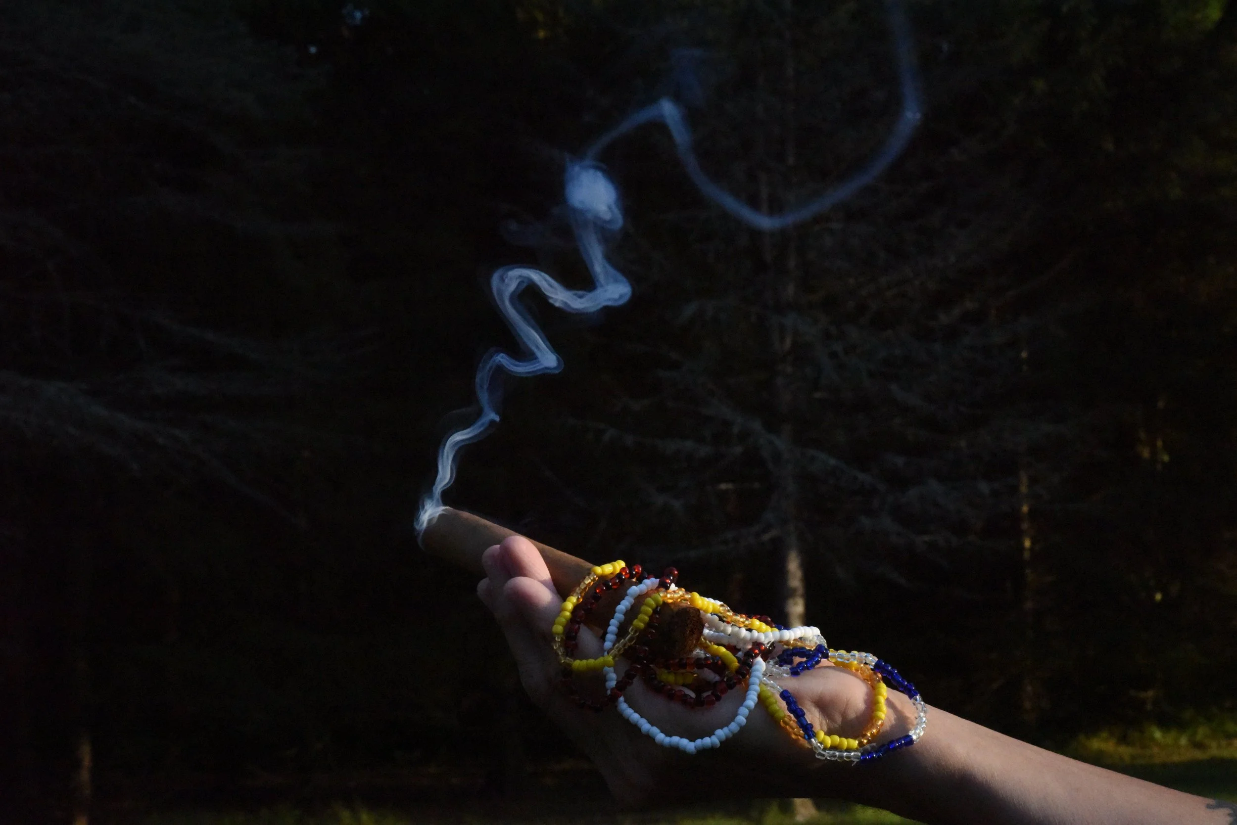 A hand with colorful beaded bracelets holding a lit cigarette with smoke rising into a dark night background.