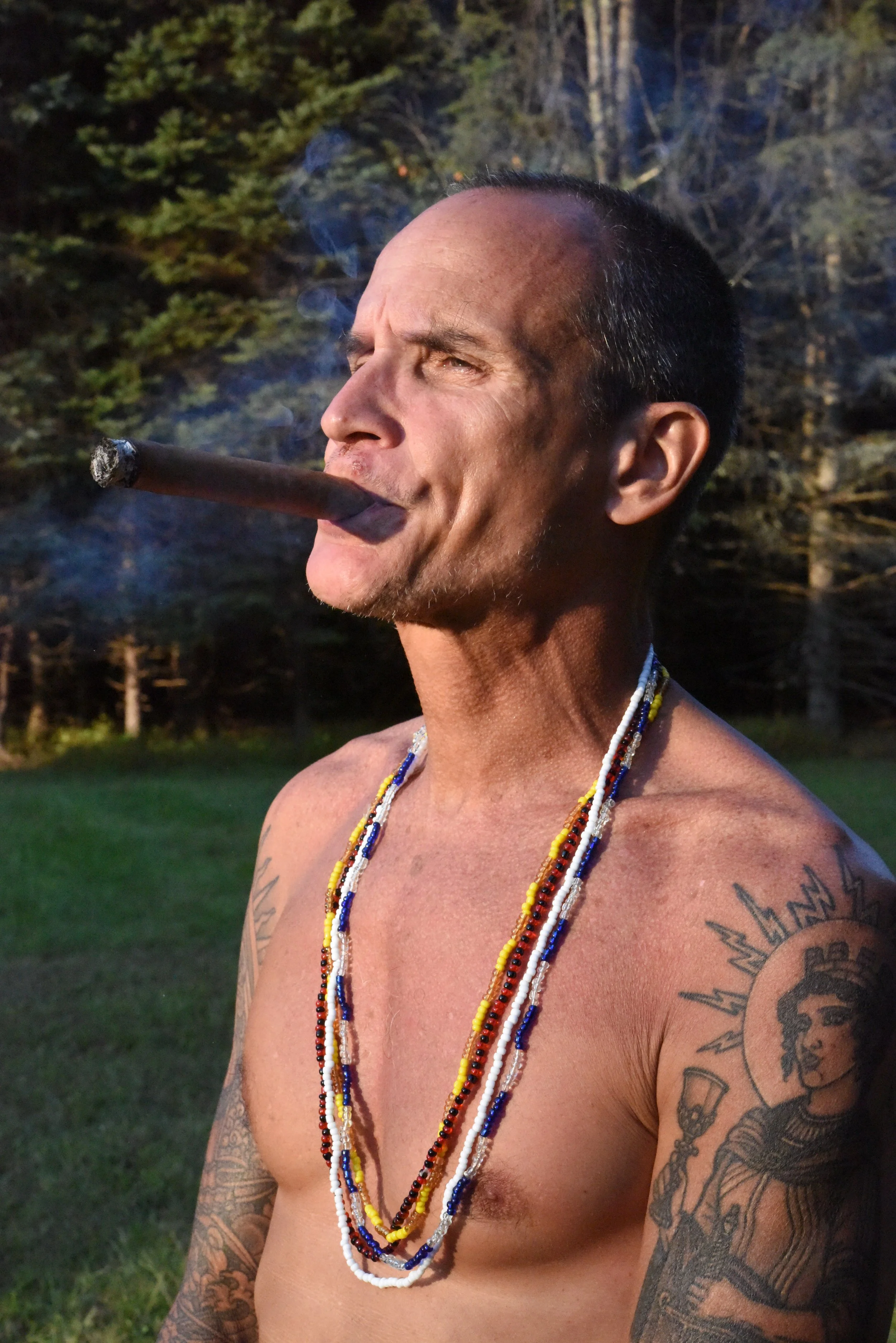 A shirtless man with tattoos and multicolored beaded necklaces, smoking a cigar outdoors in a natural setting with trees in the background.