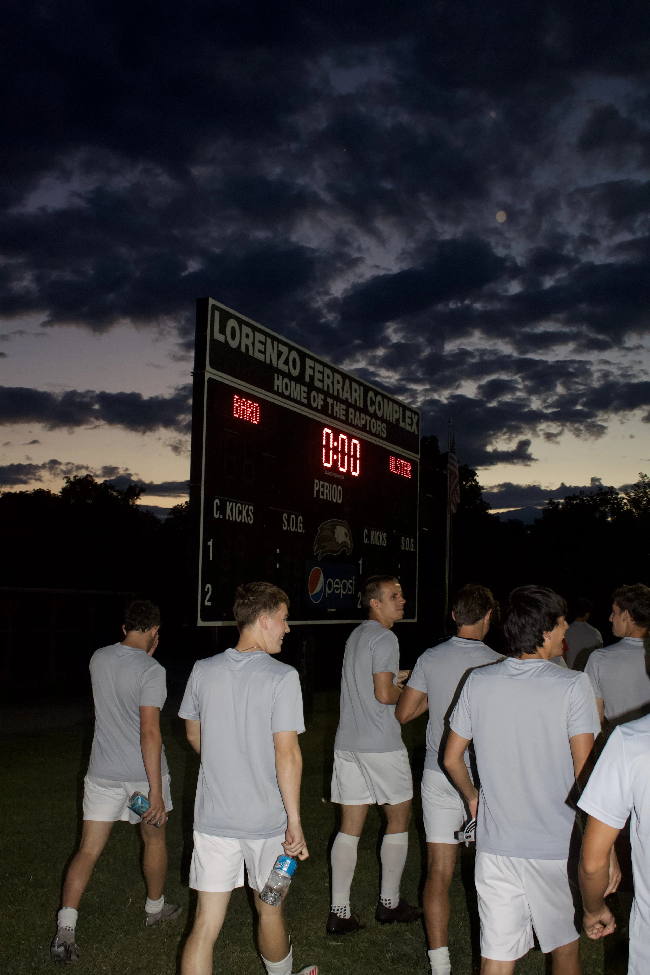 Soccer players in white uniforms standing on a field after a game at Lorenzo Ferrari Complex, with a dark evening sky with clouds and a visible moon, scoreboard showing zeroes, and spectators in the background.