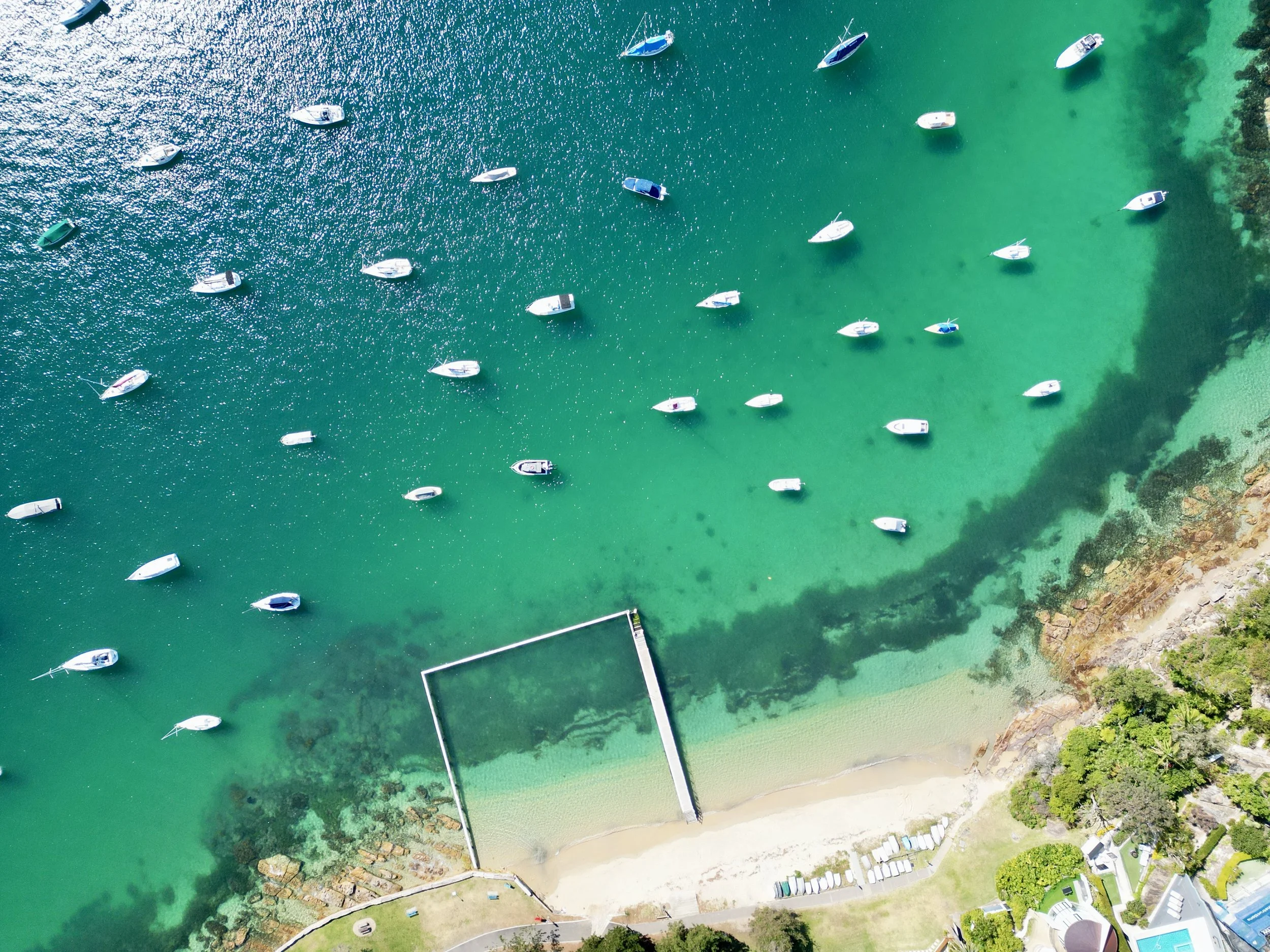 An aerial view of a marina with numerous boats anchored in clear, greenish water near a small beach area and rocky shoreline, with some greenery and buildings nearby.