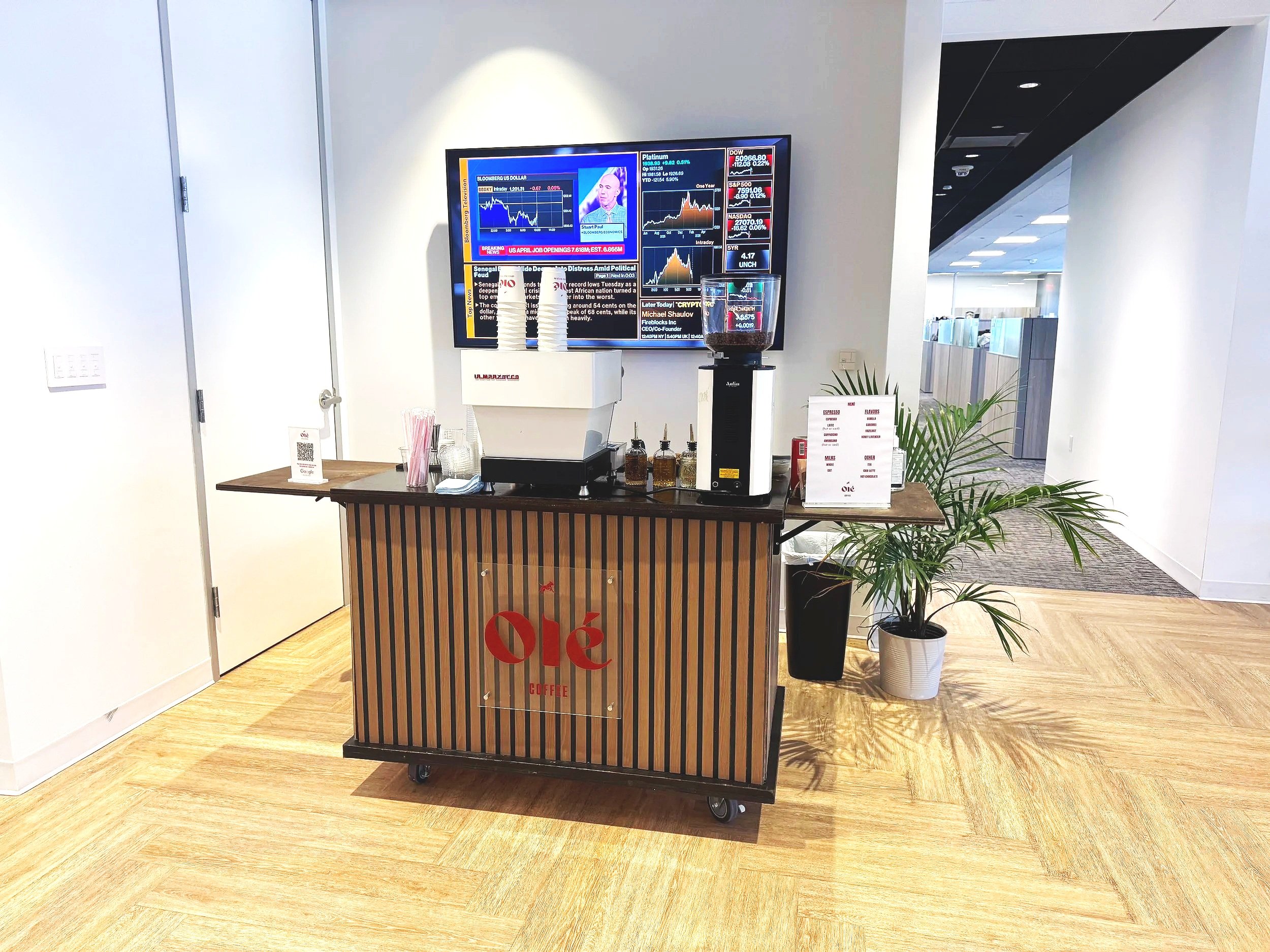 A coffee stand with a black and wood exterior that displays the word "Øle" in red letters. Behind the stand, there is a large flat-screen TV mounted on the wall showing financial news and stock market graphs, with a glass jar of coffee beans on top of the stand. To the right, there is a potted plant, and in the background, a hallway with offices or rooms.
