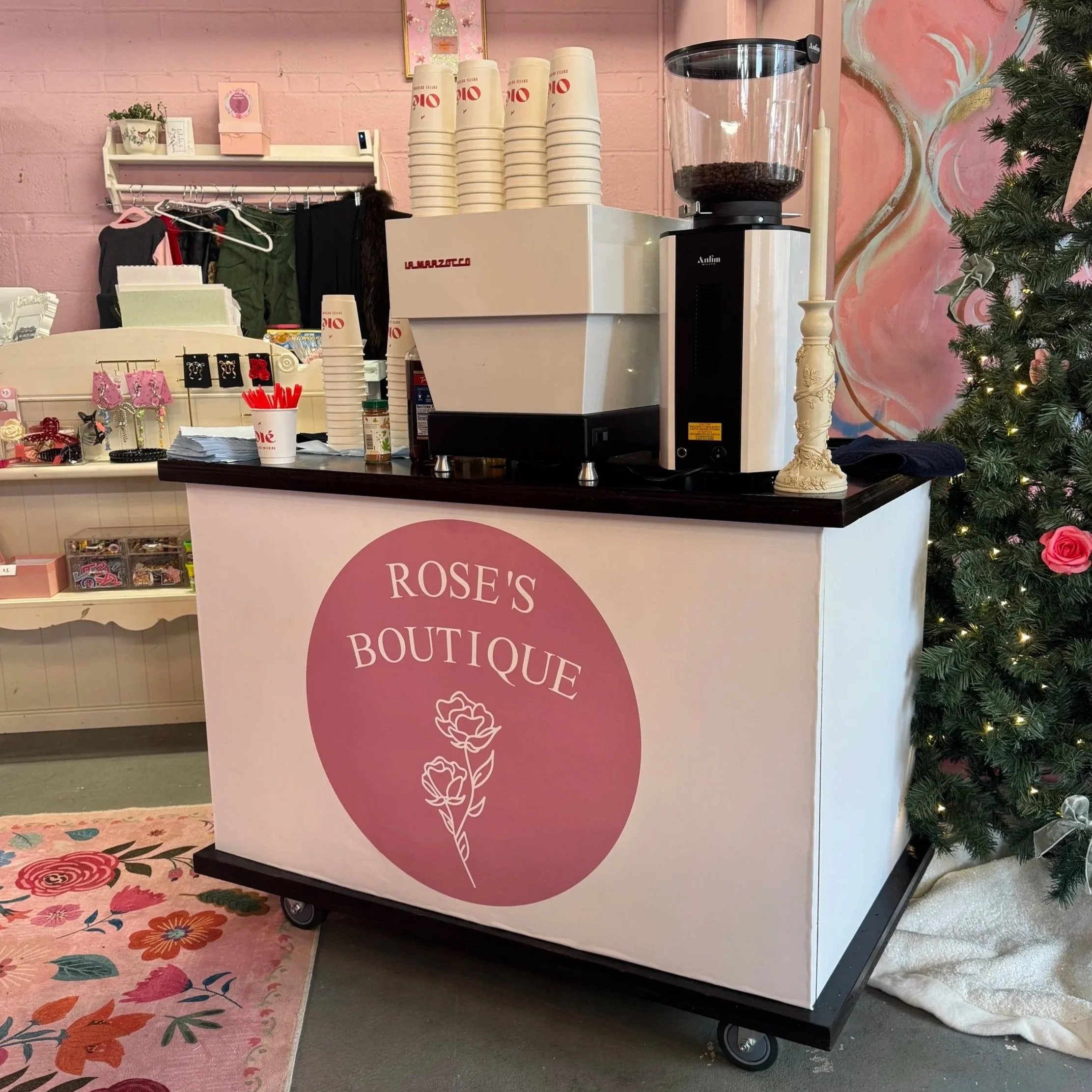 A pink and white coffee counter with the sign 'Rose's Boutique', featuring a rose illustration, espresso machinenery, next to a decorated Christmas tree with lights and pink roses.