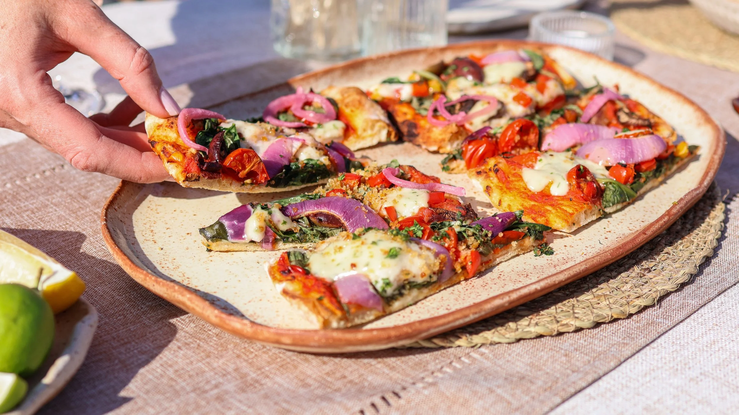 speckled ceramic platter, with pizza slices that have cheery tomatoes, red onions cheese and spinach on top and there is a hand picking up one slice of pizza in the bottom left corner