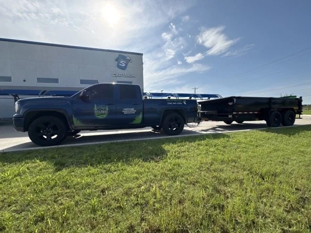 A black pickup truck parked on a grassy area next to a building, pulling a trailer. The building has a logo that resembles a blue shield with text, and the truck has decals and equipment on the bed.