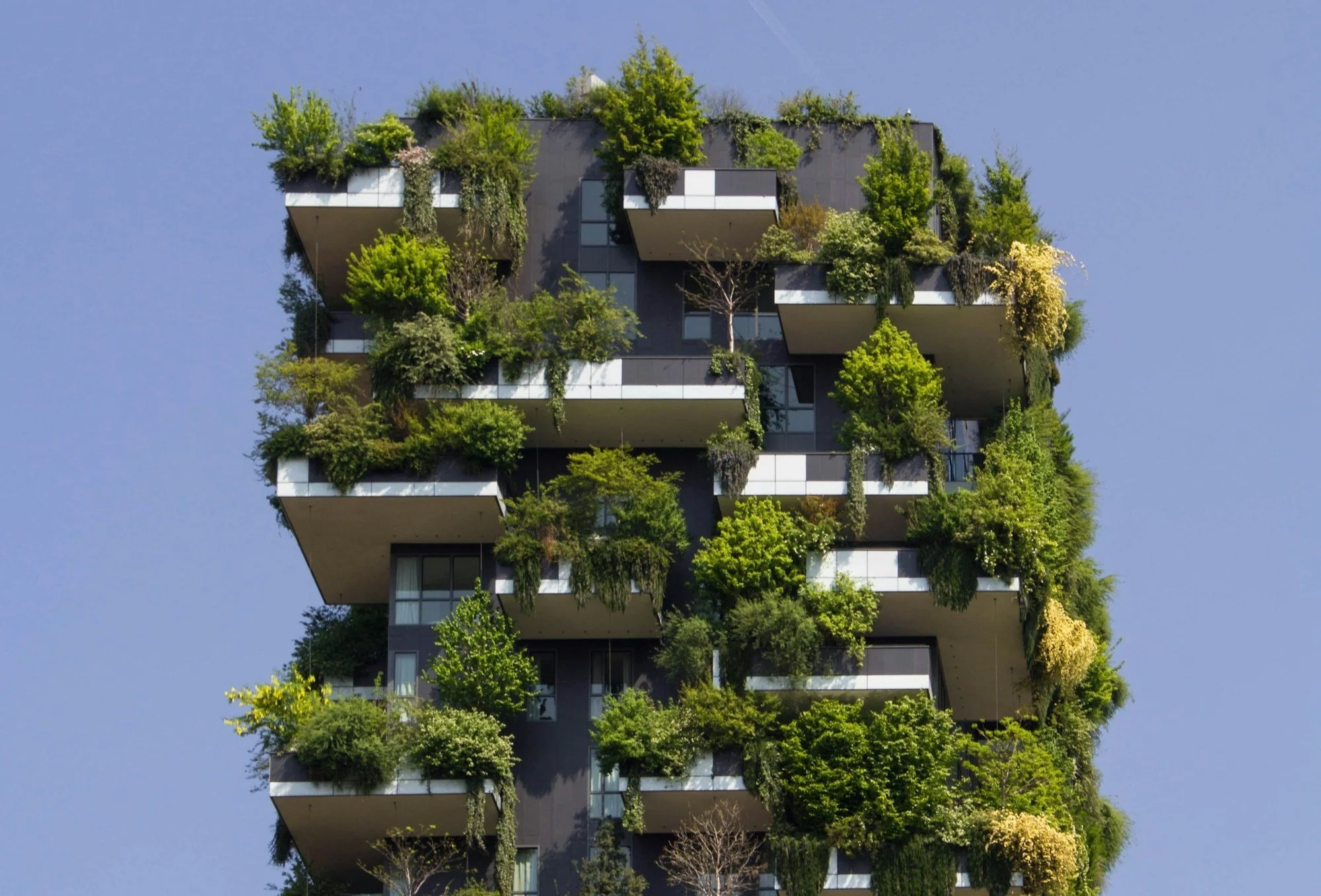 A modern multi-story building with numerous balconies filled with lush green trees, plants, and shrubs against a clear blue sky.