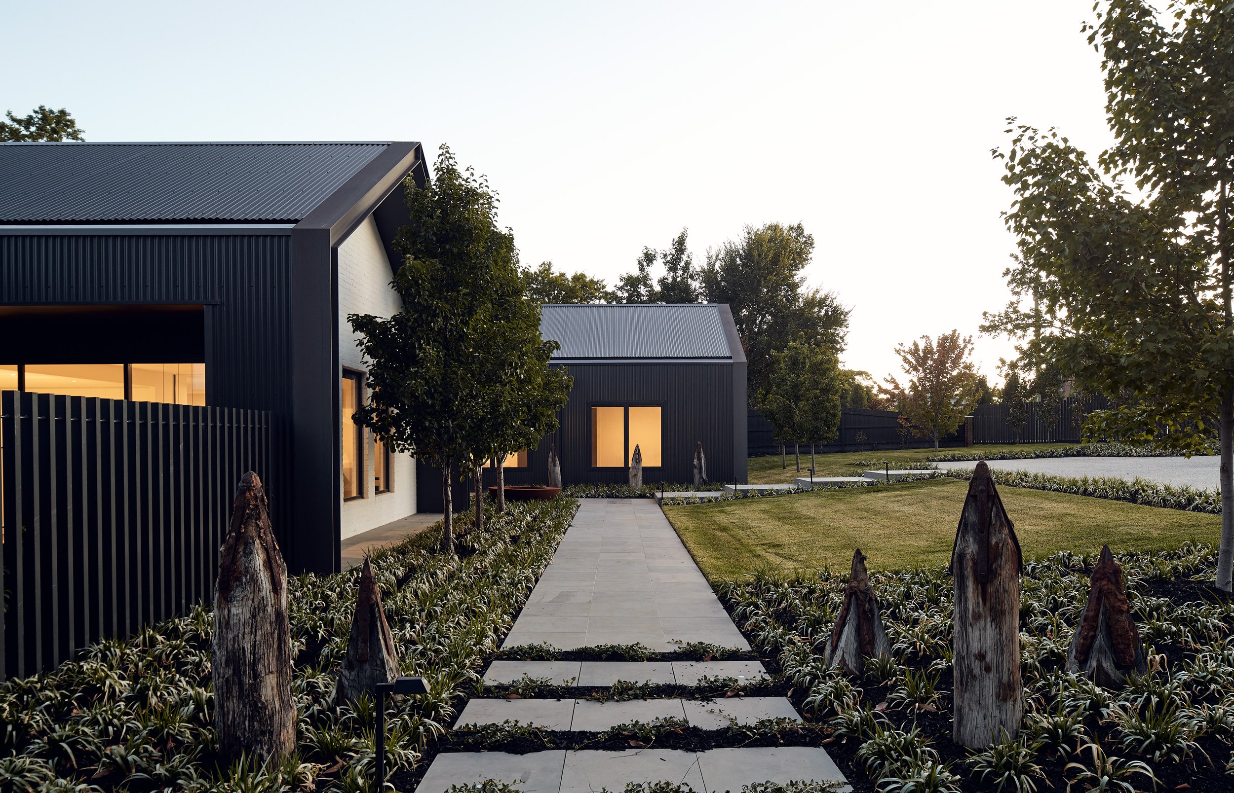 Modern house with black exterior and metal roof, lit windows, surrounded by a well-maintained garden and trees, during early evening.