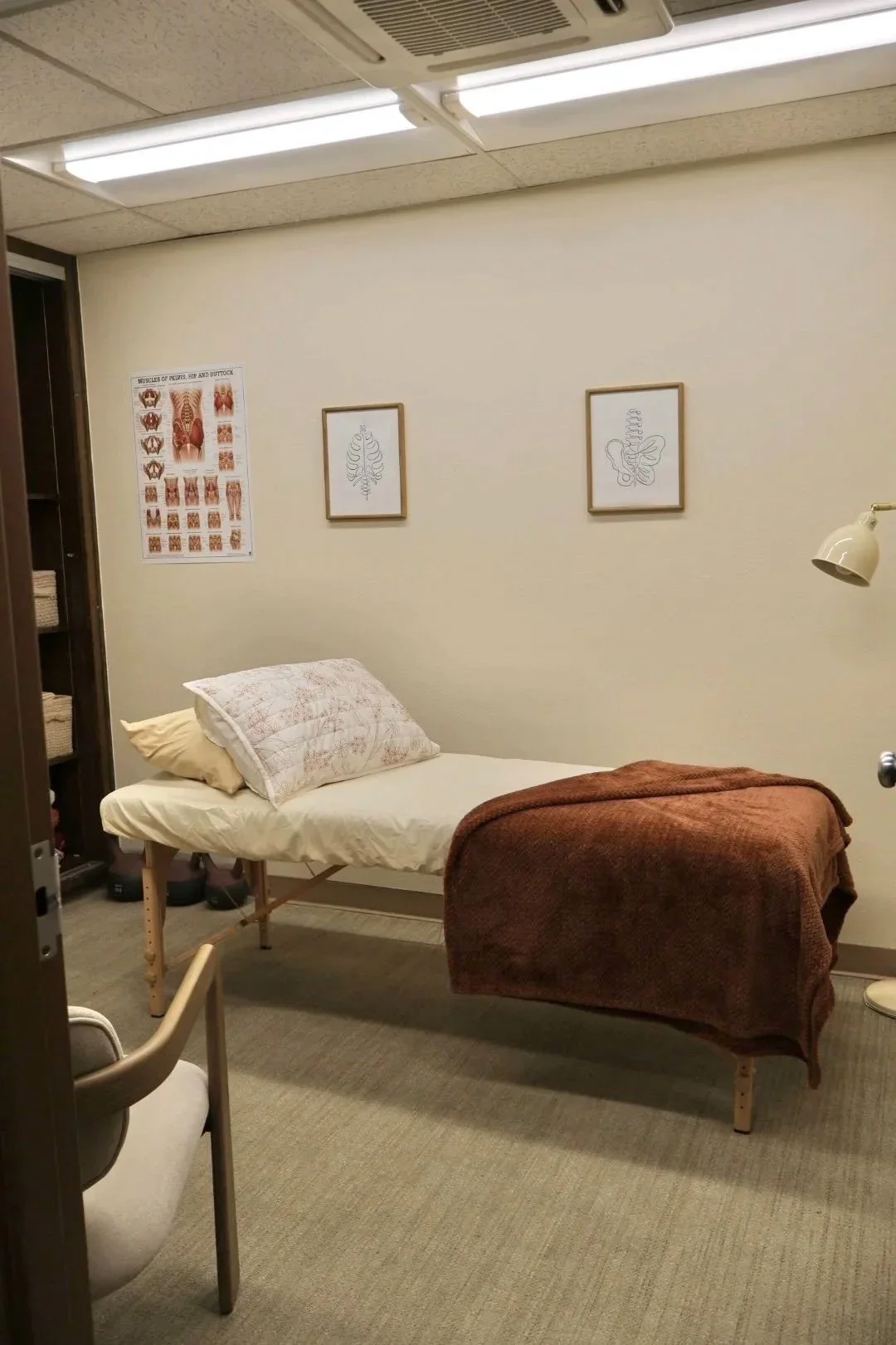 A small medical examination room with a bed covered partly with a rust-colored blanket and pillows, a wall-mounted light to the right, a beige chair in front of the bed, and framed anatomical sketches on the wall. There is a poster with muscular diagrams on the left wall.