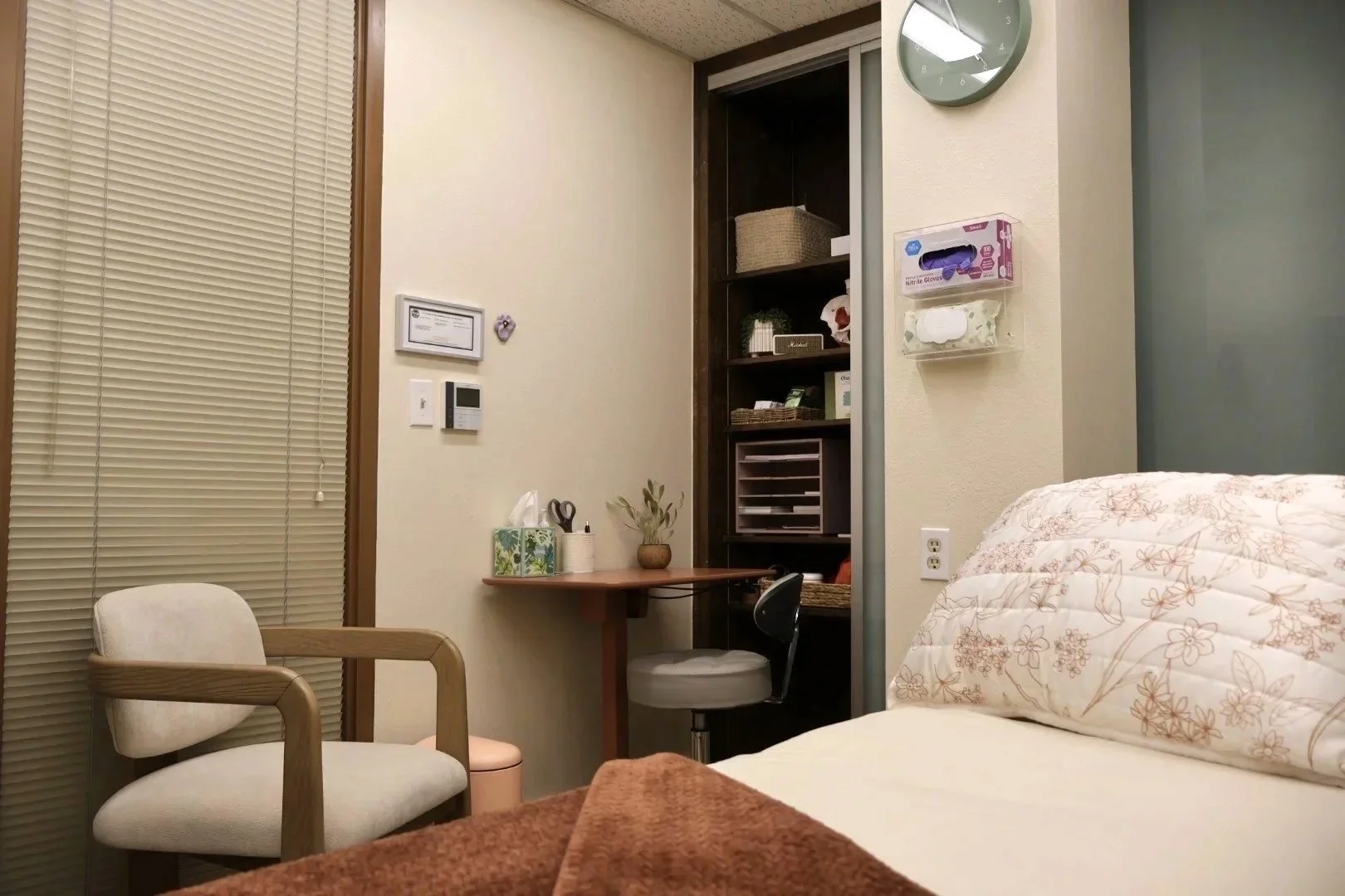 A cozy bedroom corner with a bed, a white cushioned armchair, a small desk with a chair, and a sliding closet door with shelves inside. The wall has a clock, tissue box, and some decorative items.