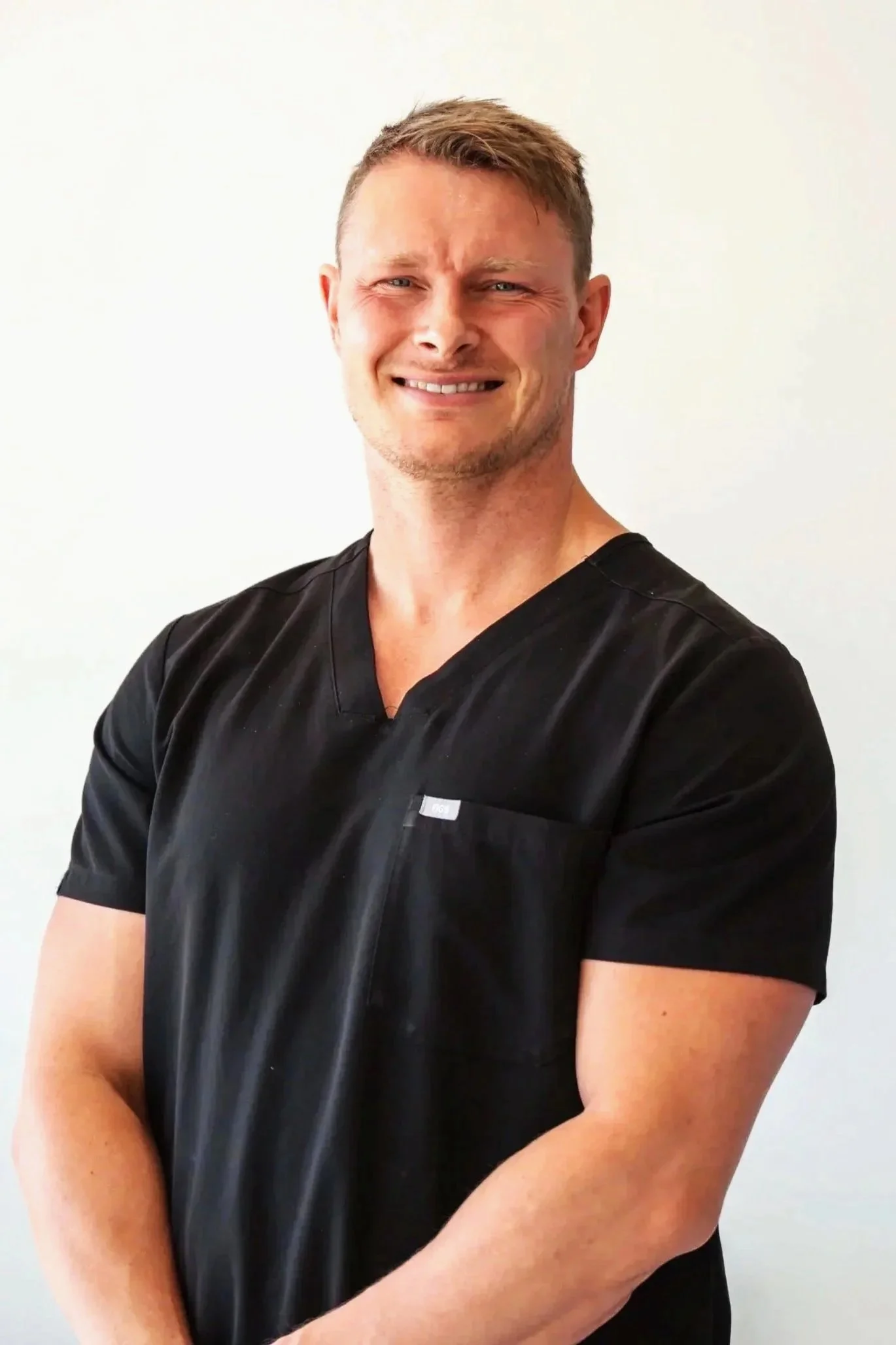 A smiling man with short light brown hair, wearing a black V-neck shirt, standing against a green wall.