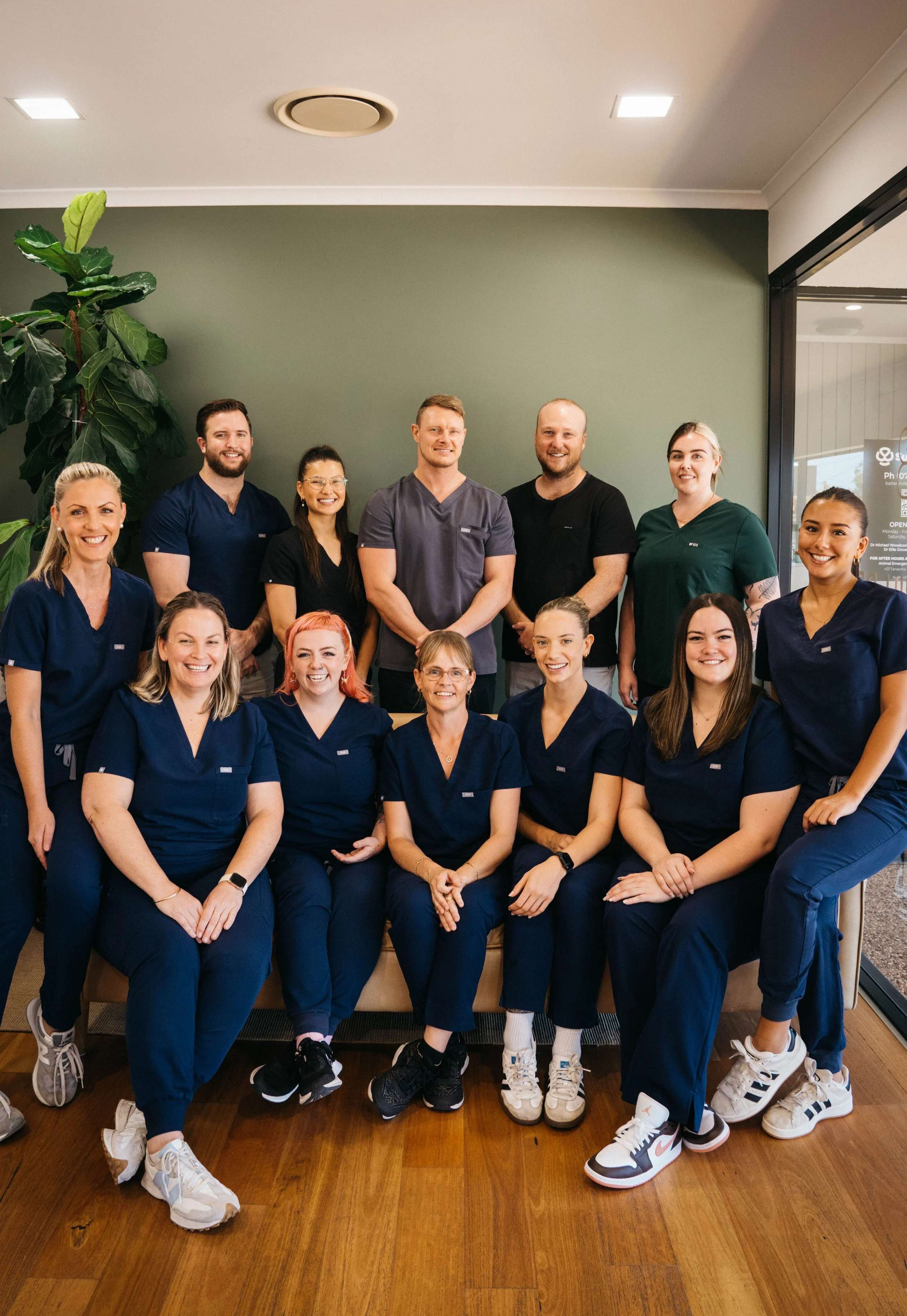 Group photo of healthcare professionals, including nurses and staff, standing and sitting indoors against a green wall, smiling at the camera.