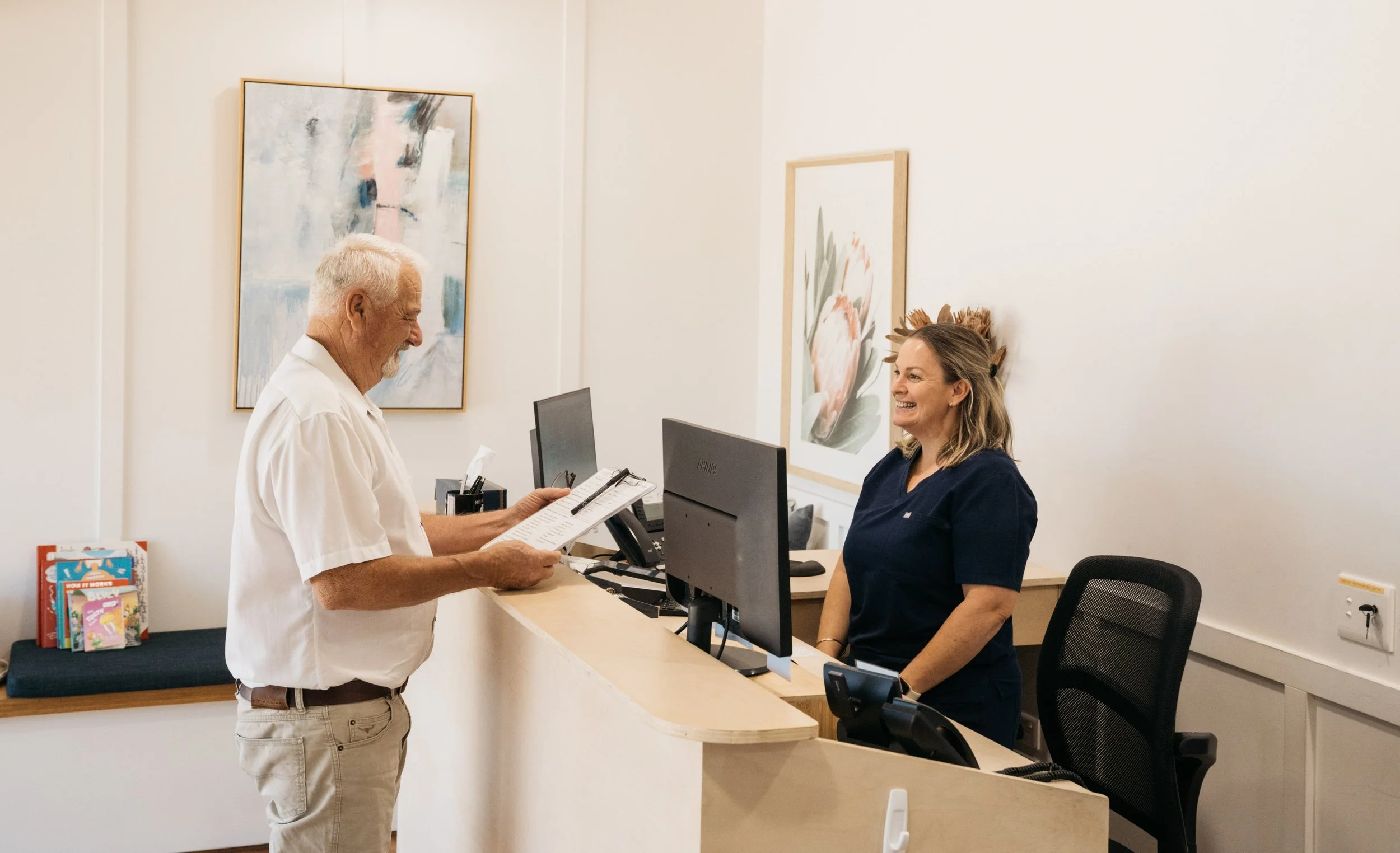 An elderly man interacts with a smiling female receptionist at a medical front desk, with artwork on the wall behind them.