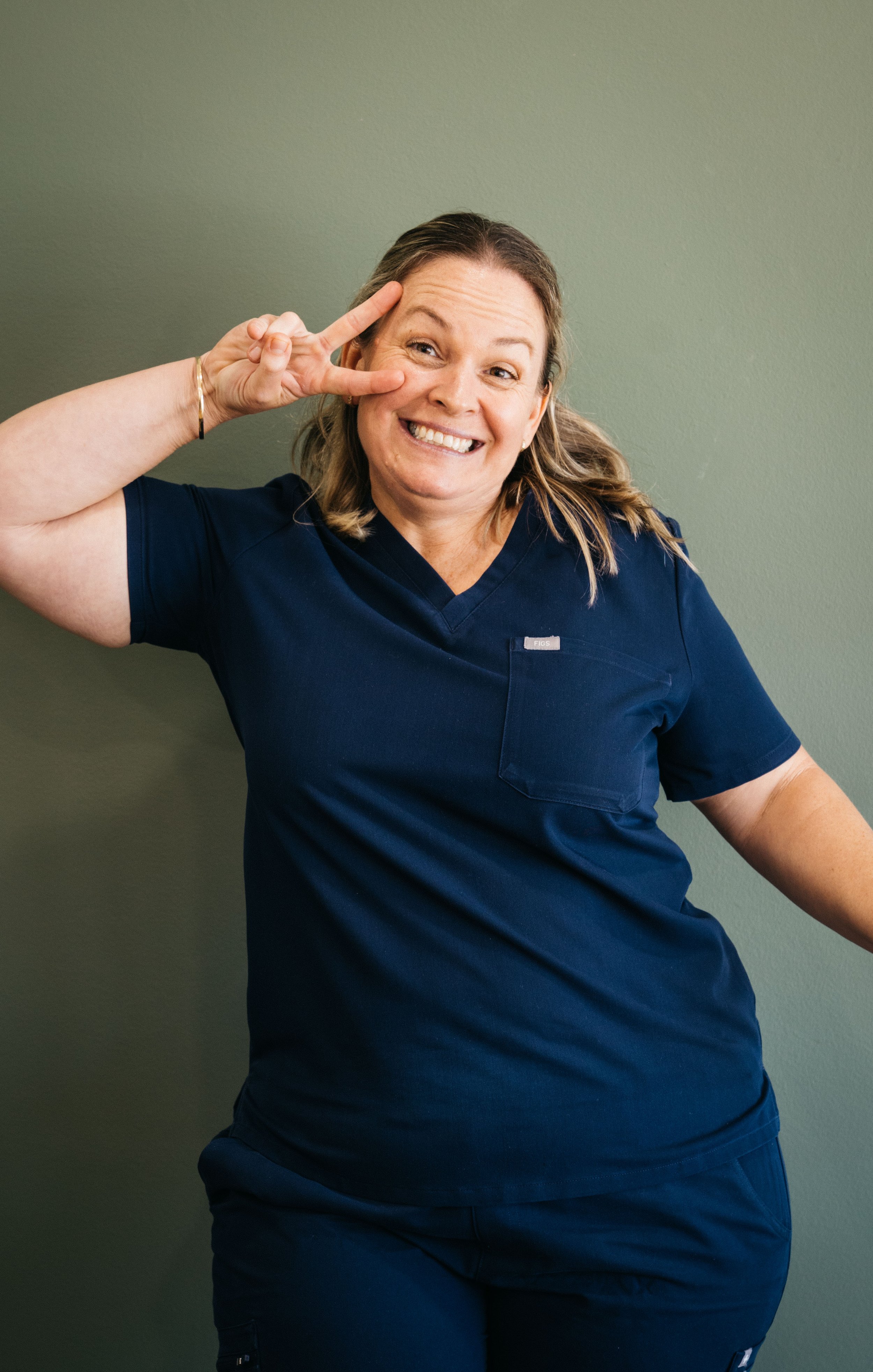 A woman in navy scrubs smiling and making a peace sign near her eye against a green wall.