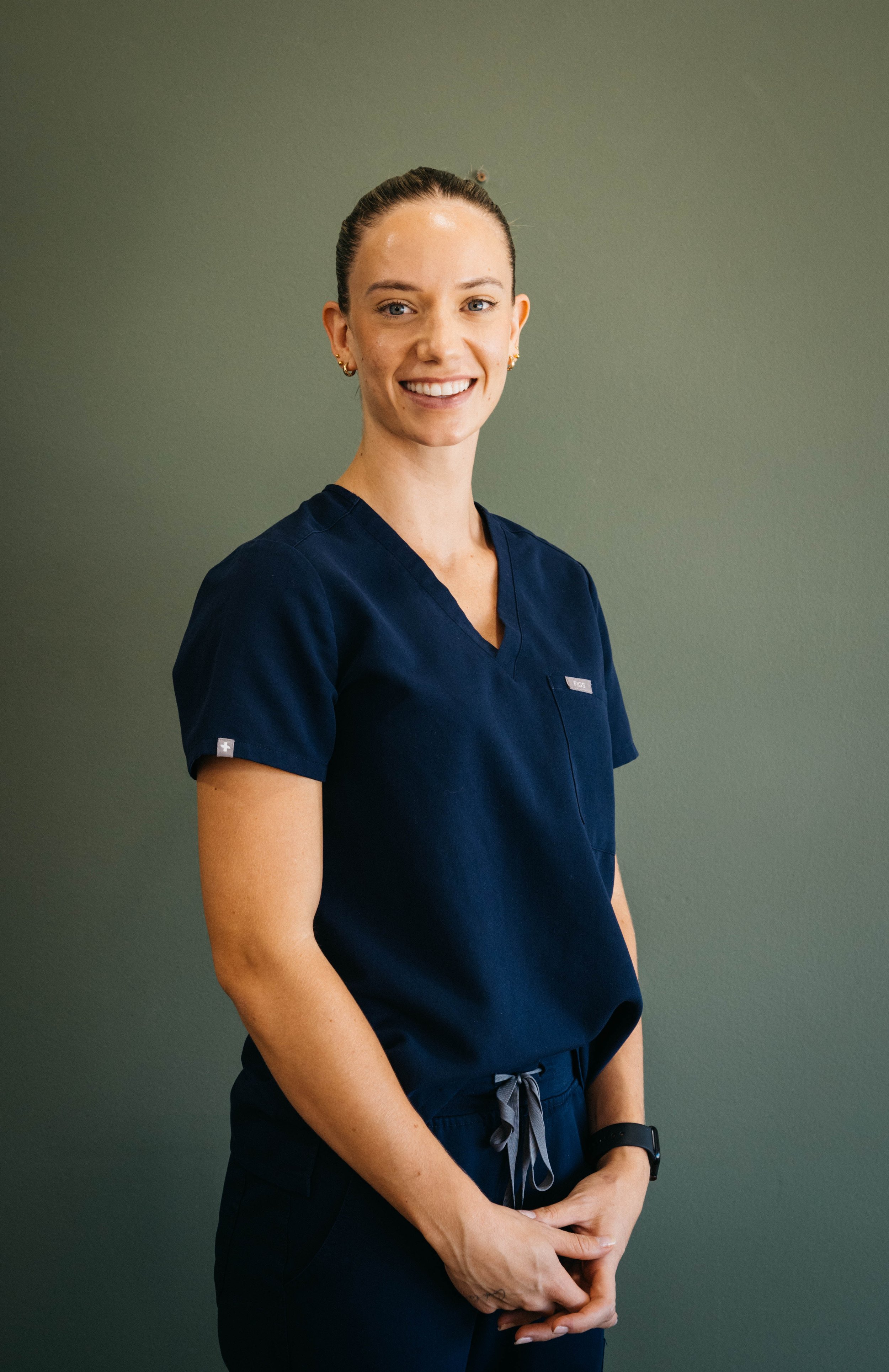 A smiling woman in navy medical scrubs standing against a gray wall.