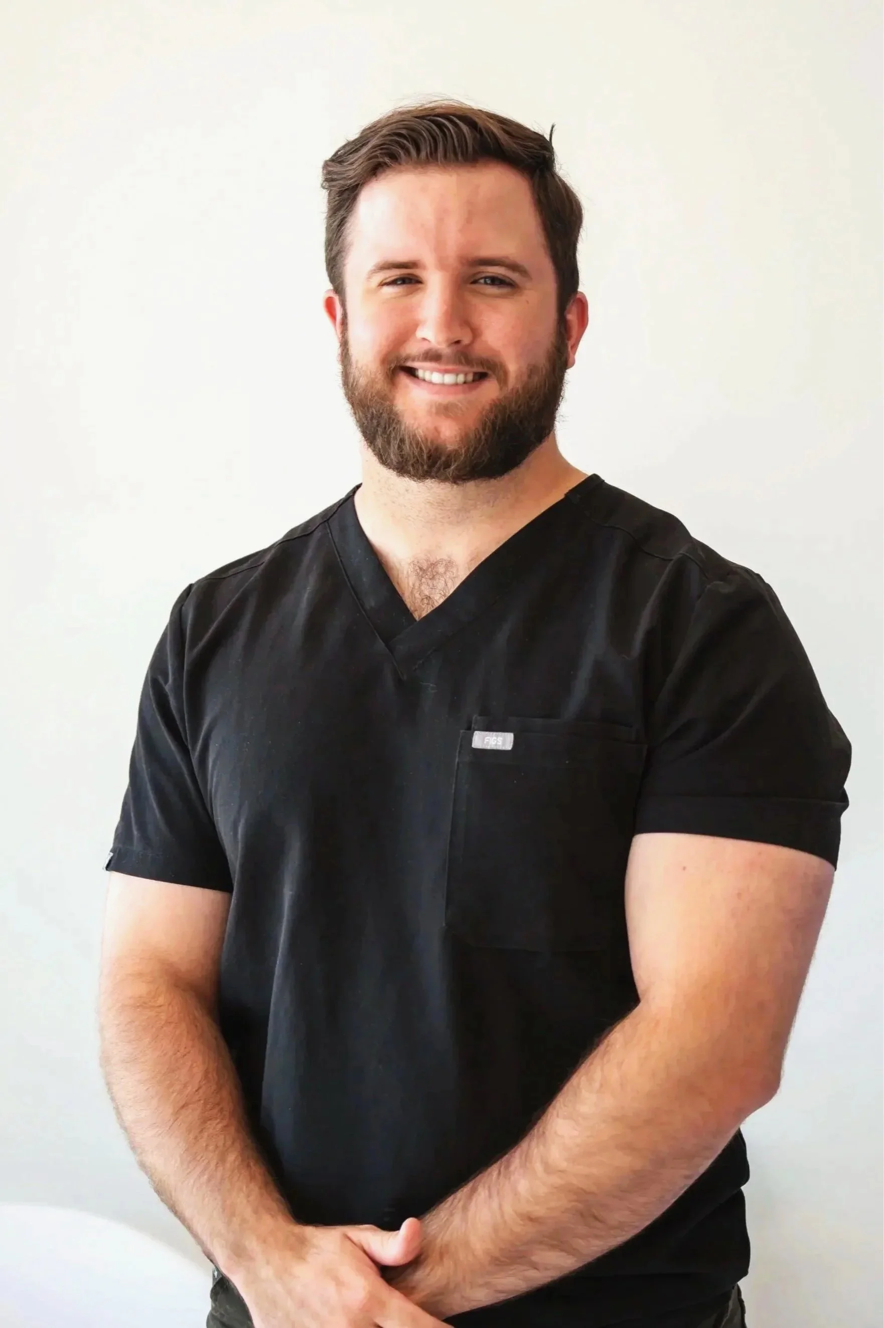 A young man with brown hair, beard, and blue eyes smiling, wearing a navy blue polo shirt with a small label on the chest, standing against a gray background.