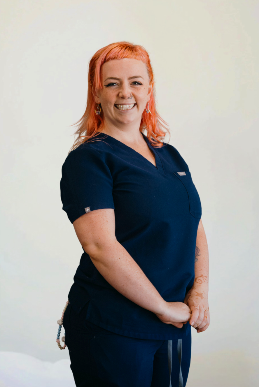A smiling woman with pink hair, tattoos, and earrings wearing navy scrubs stands against a plain green wall.
