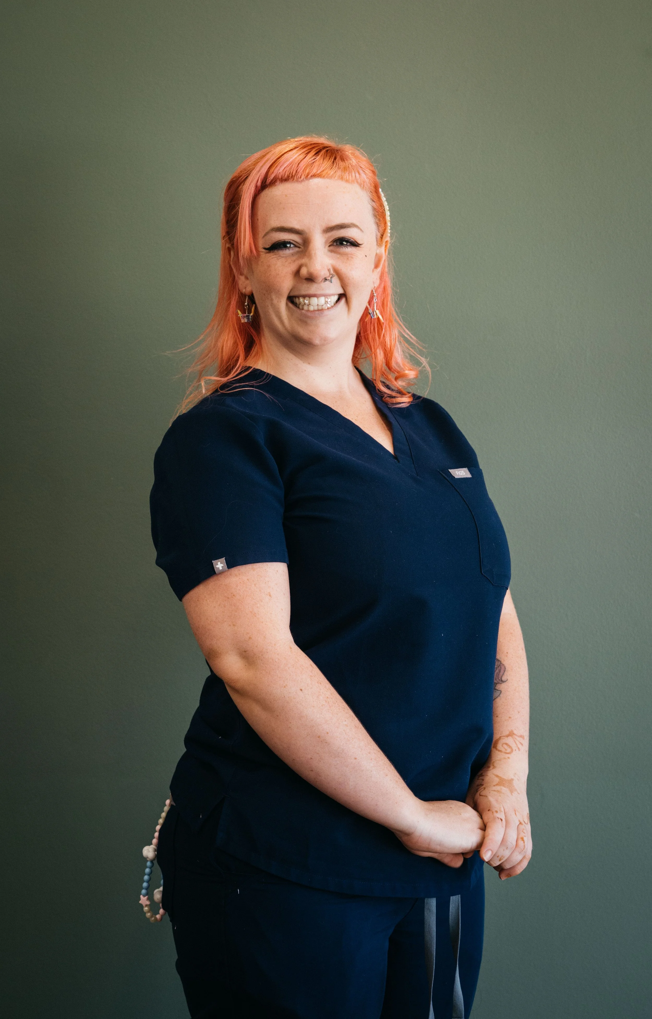 A smiling woman with pink hair, tattoos, and earrings wearing navy scrubs stands against a plain green wall.