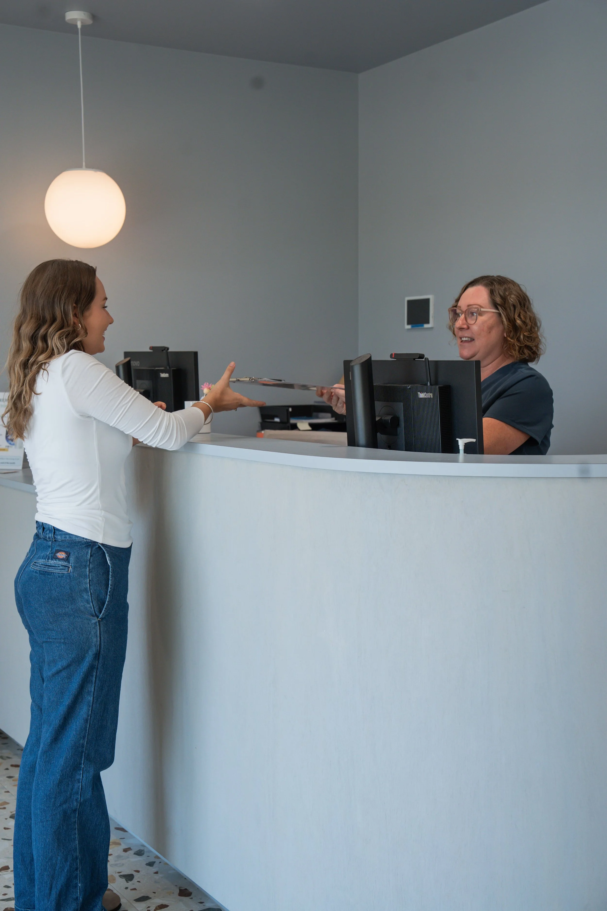 An elderly man interacts with a smiling female receptionist at a medical front desk, with artwork on the wall behind them.