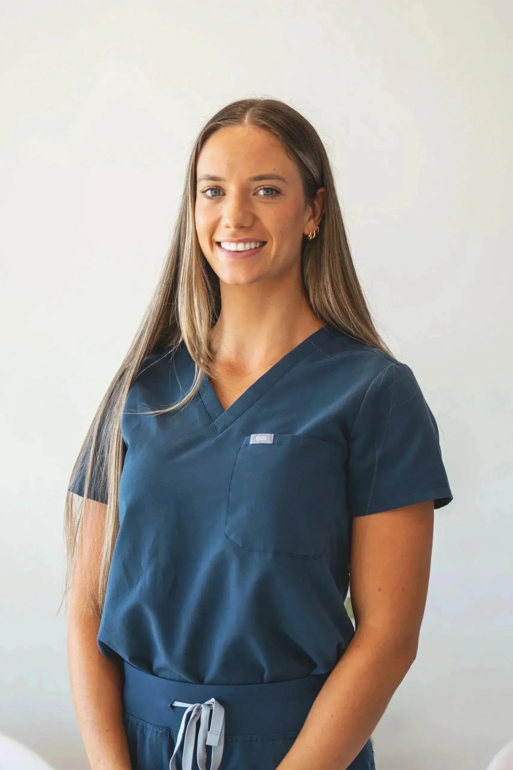 A smiling woman in navy medical scrubs standing against a gray wall.