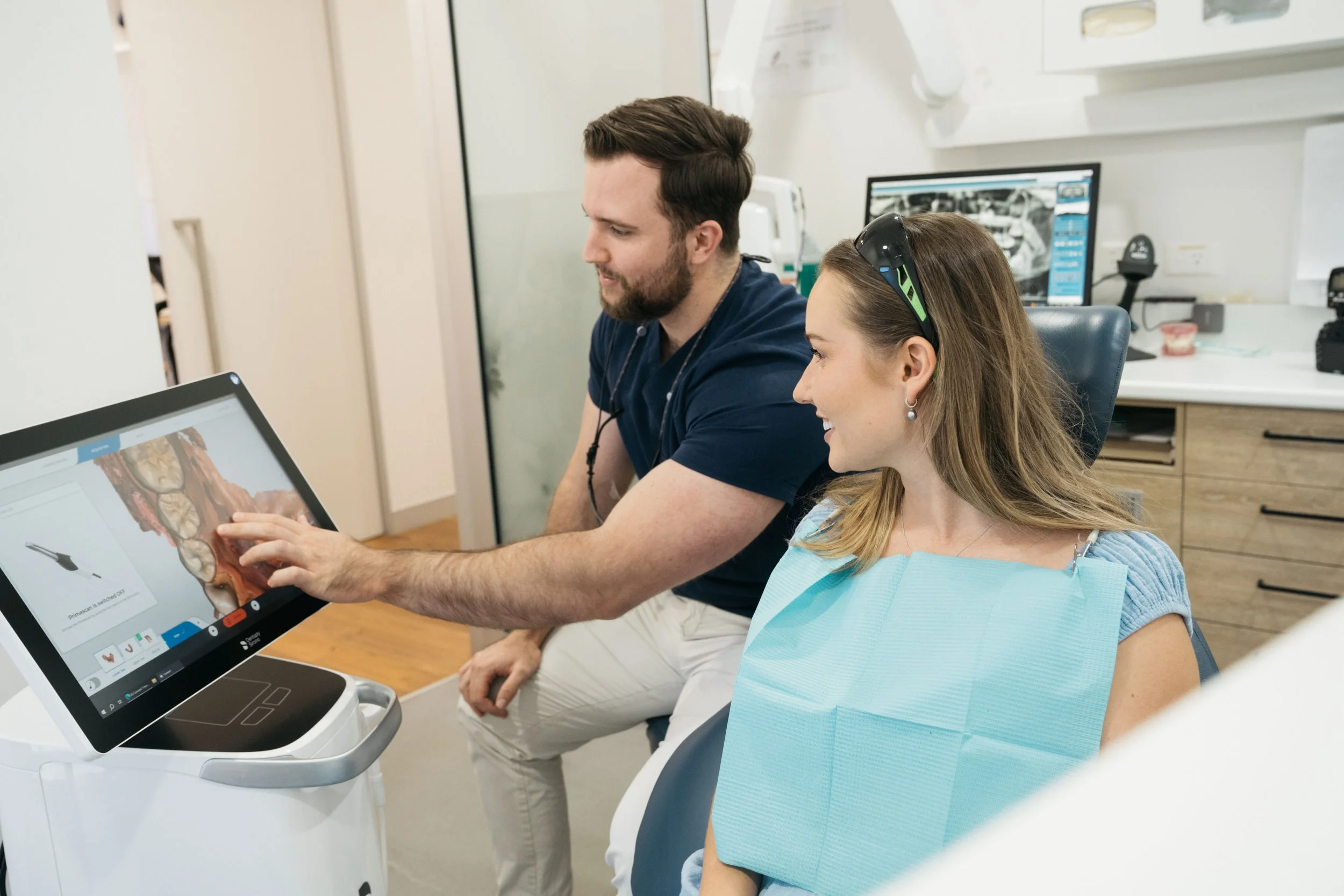 A dentist and a woman patient in a dental office looking at a computer screen showing a dental scan or X-ray.