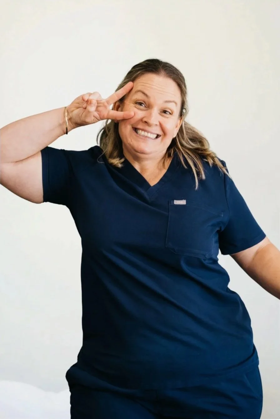 A woman in navy scrubs smiling and making a peace sign near her eye against a green wall.