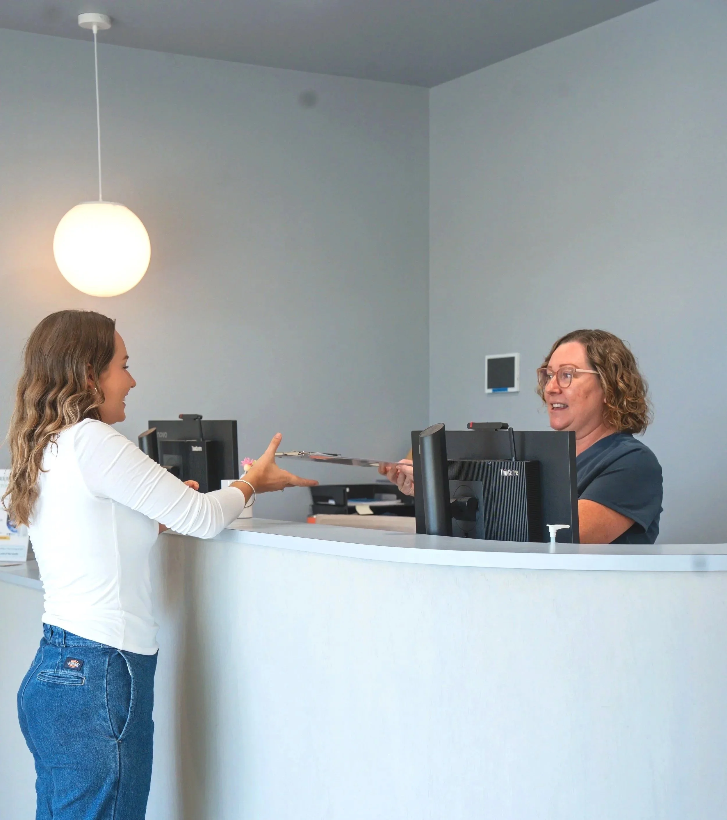 An elderly man interacts with a smiling female receptionist at a medical front desk, with artwork on the wall behind them.
