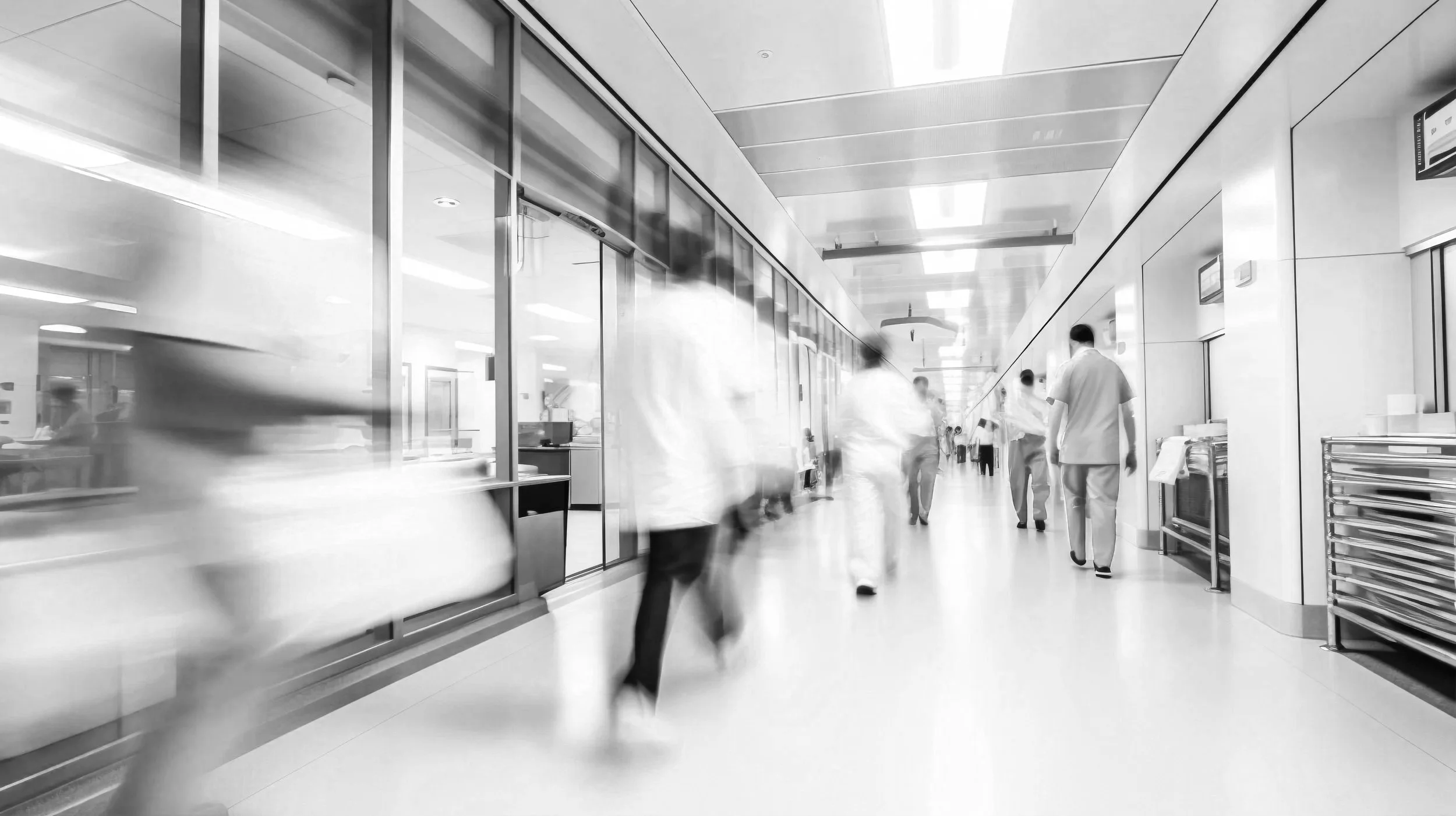 Hospital corridor with blurred medical staff and visitors walking, bright lighting, and clinical surroundings.