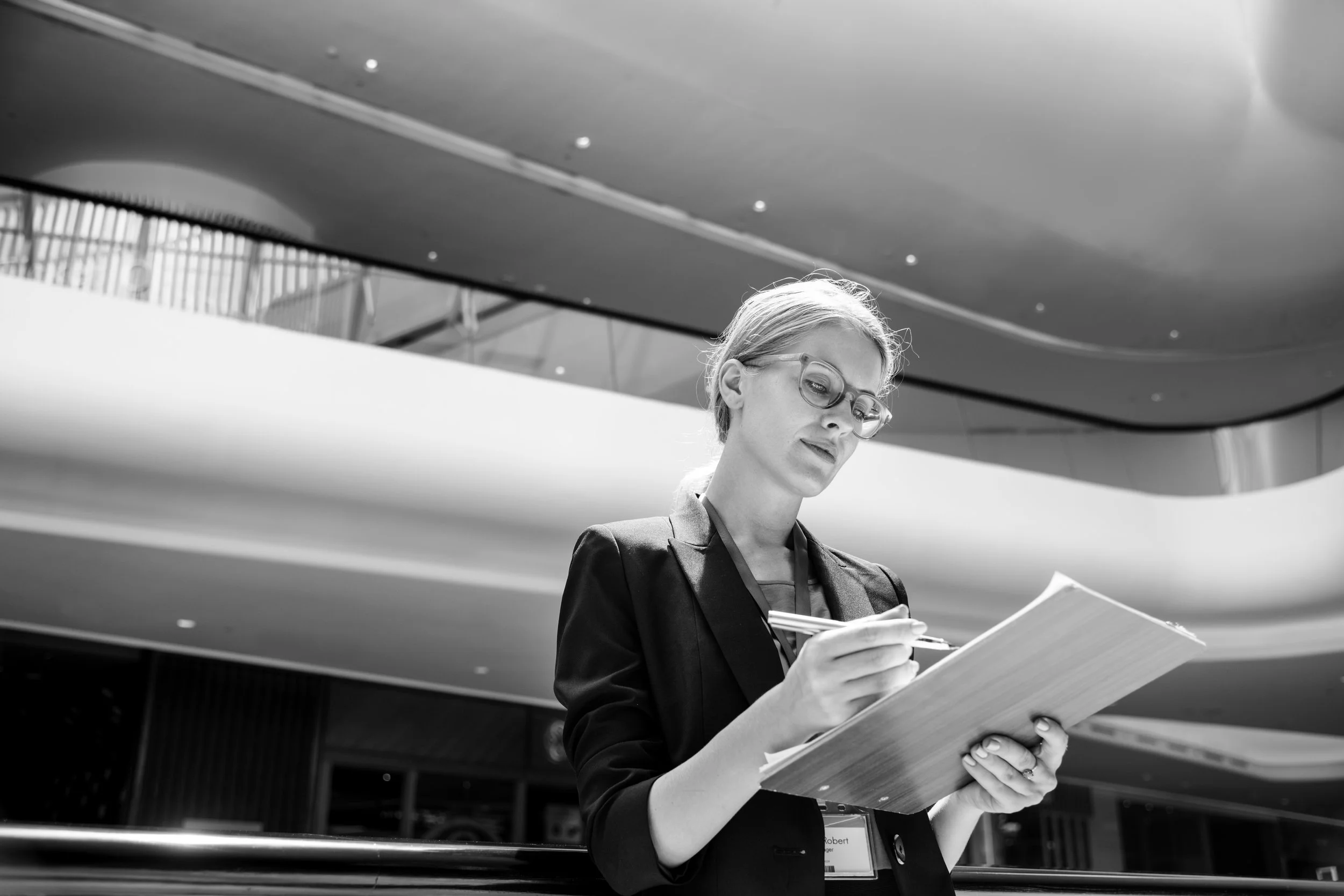 Black and white photo of a woman with glasses writing on a notepad in a modern indoor space, possibly a conference or office building.