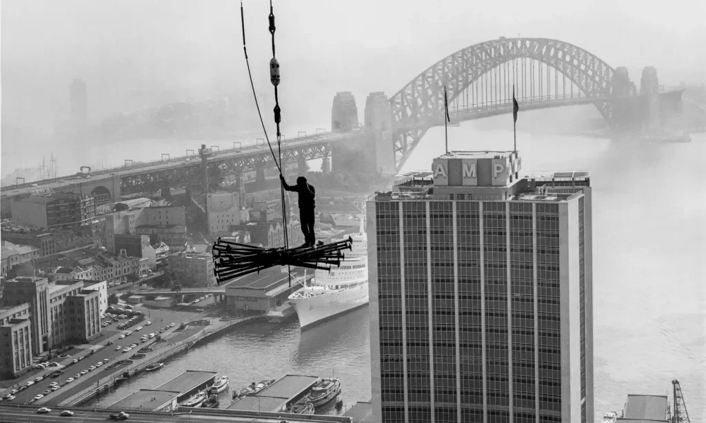 A person working on a high wire suspended between two tall buildings in an urban cityscape, with a large river and a famous arch bridge in the background.