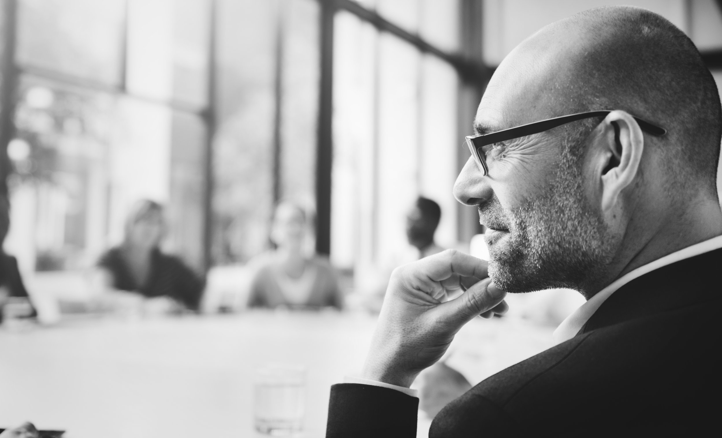 A man with glasses and a beard smiling thoughtfully while sitting at a table in a bright room with large windows, with blurred people in the background.