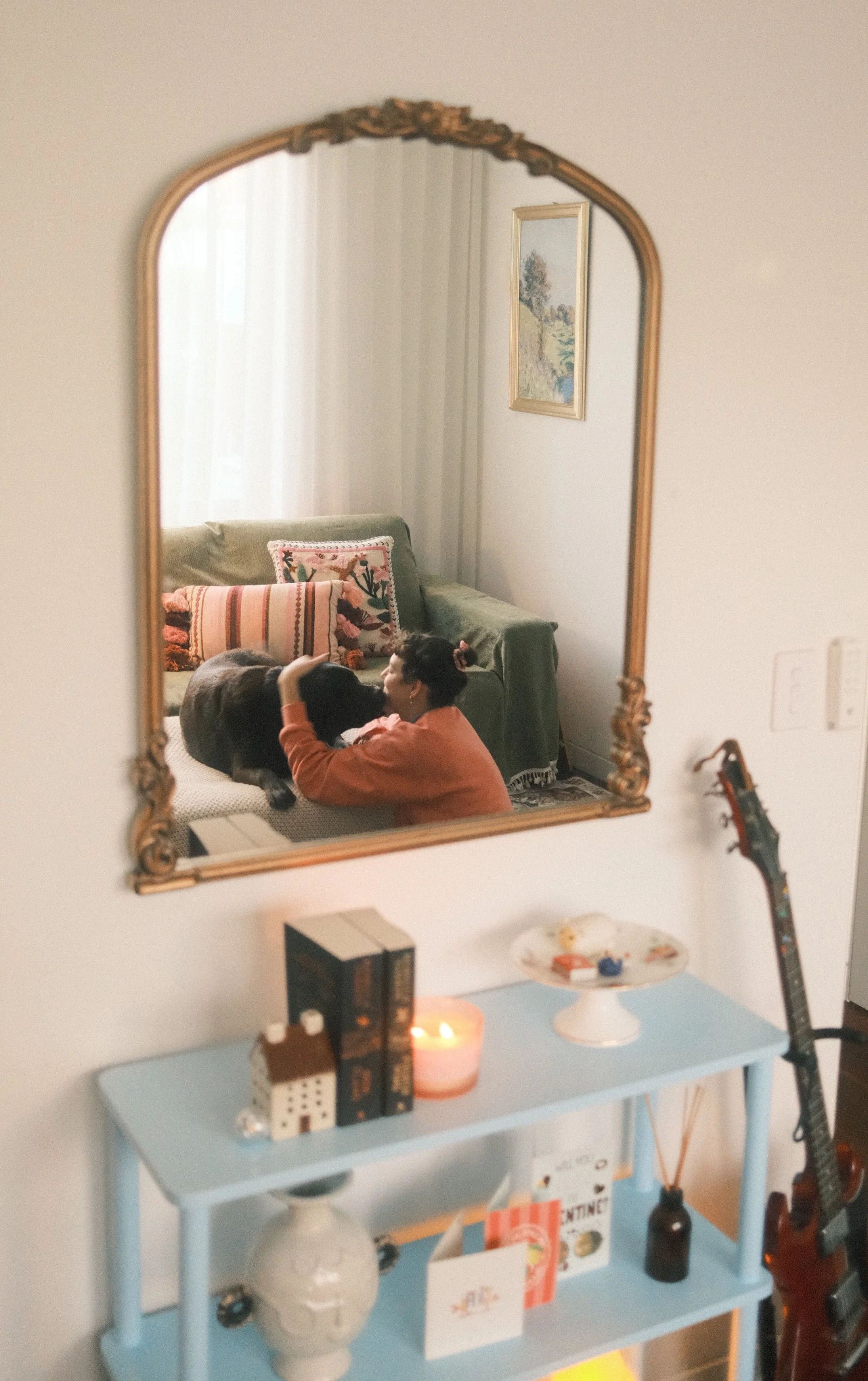 A woman is lying on a couch, leaning down to kiss or cuddle a large dog in a living room, reflected in a decorative wall mirror.