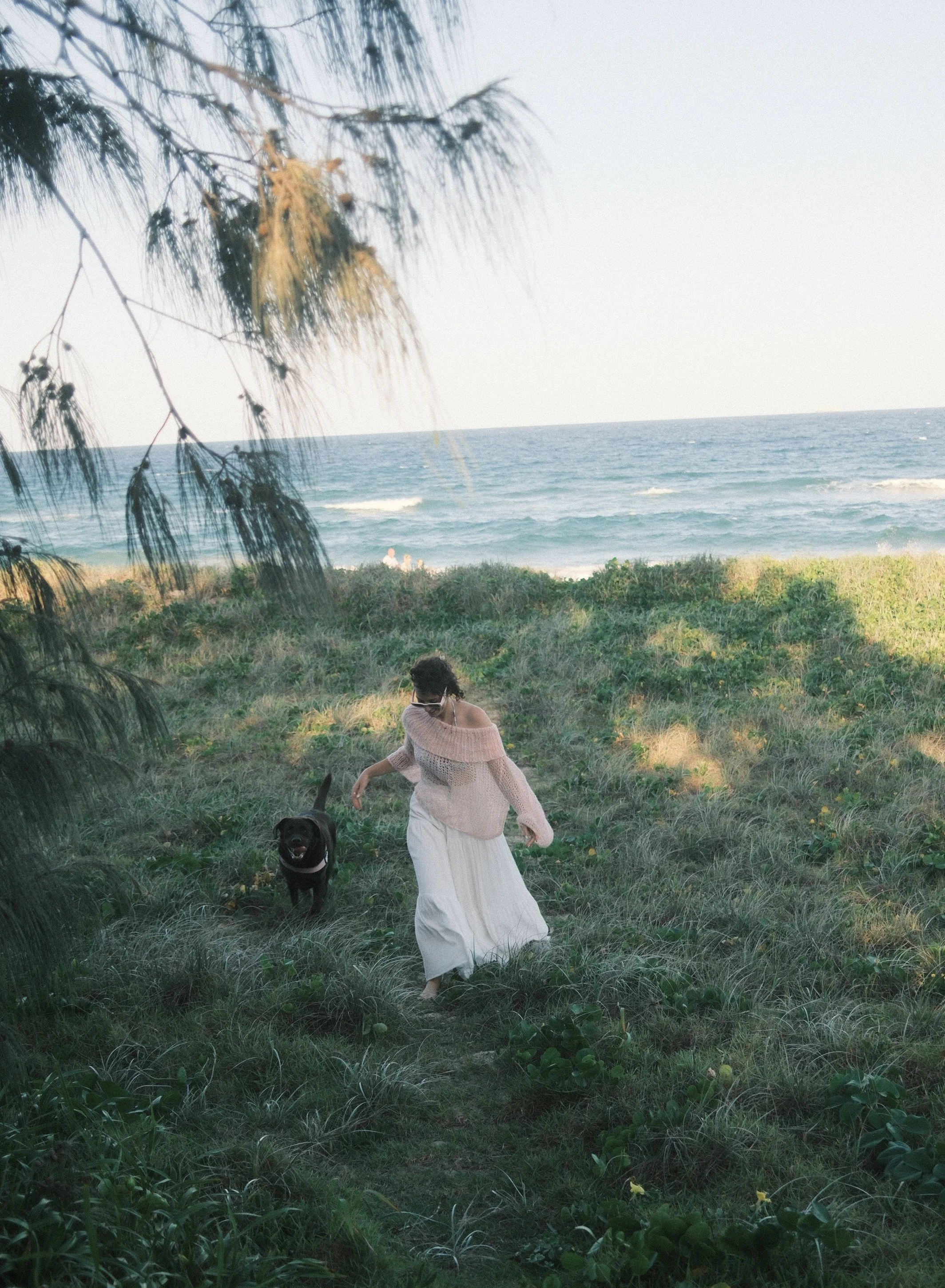 Woman in a long white skirt and pink sweater walking with a black dog on a grassy path near the beach with ocean in the background.