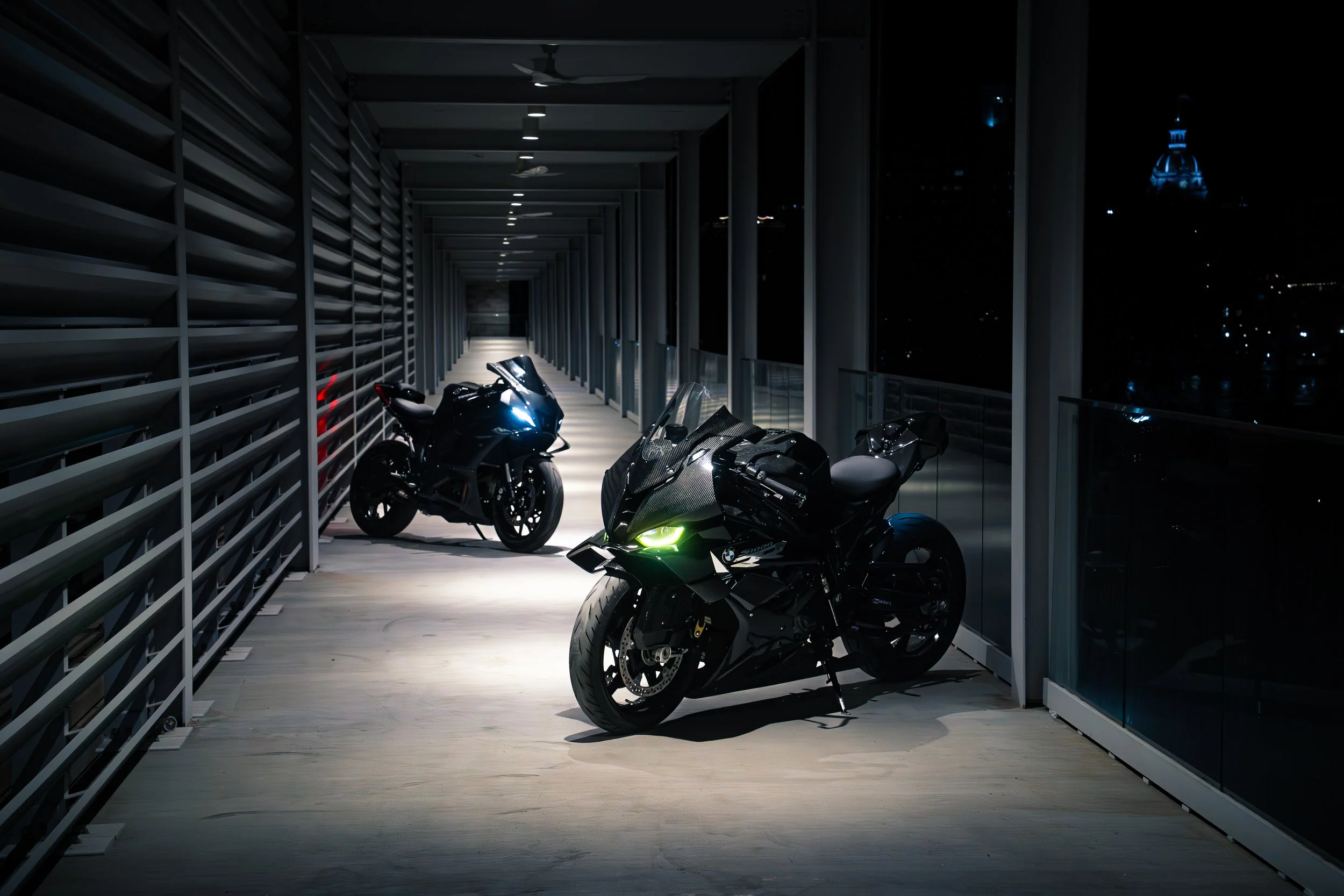Two motorcycles parked on a brightly lit balcony at night, with city lights and a domed building visible in the background.
