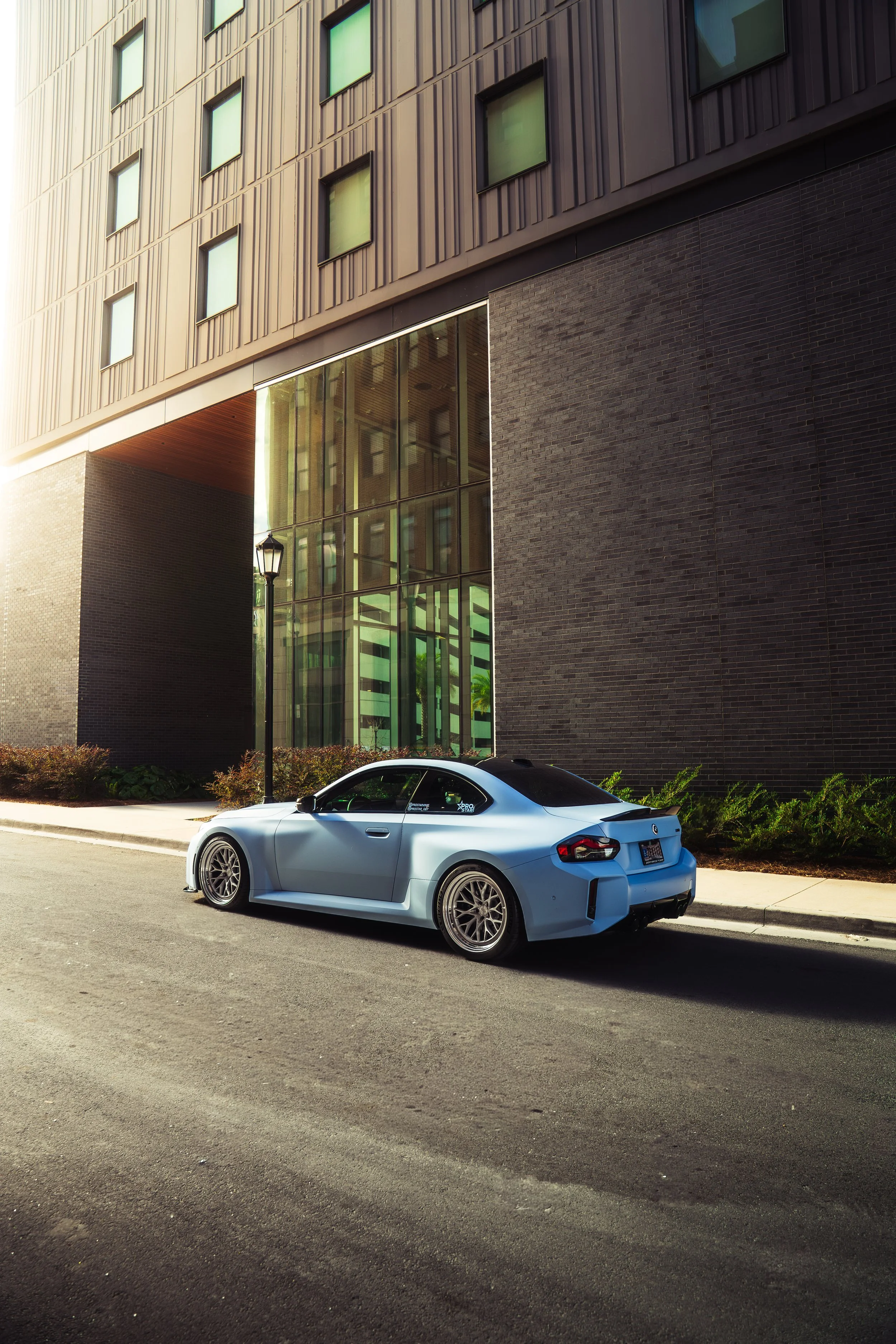 A blue sports car parked on the street in front of a modern building with glass and brick walls, sunlight casting shadows.