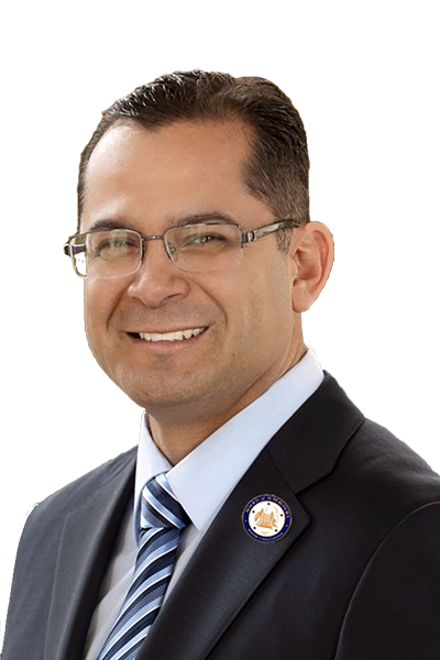 A man wearing glasses, a dark suit, and a striped tie, smiling, with a circular pin on his lapel.
