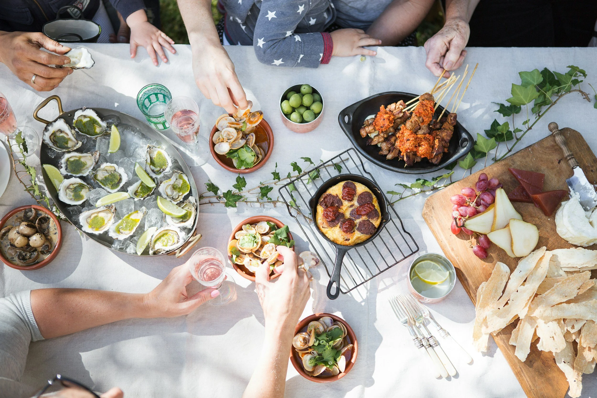 A table filled with various dishes including oysters with lemon wedges, clams, a pizza with pepperoni, grilled meat skewers, a cheese platter with fruit, and a basket of bread, with people reaching for food and holding glasses of water.
