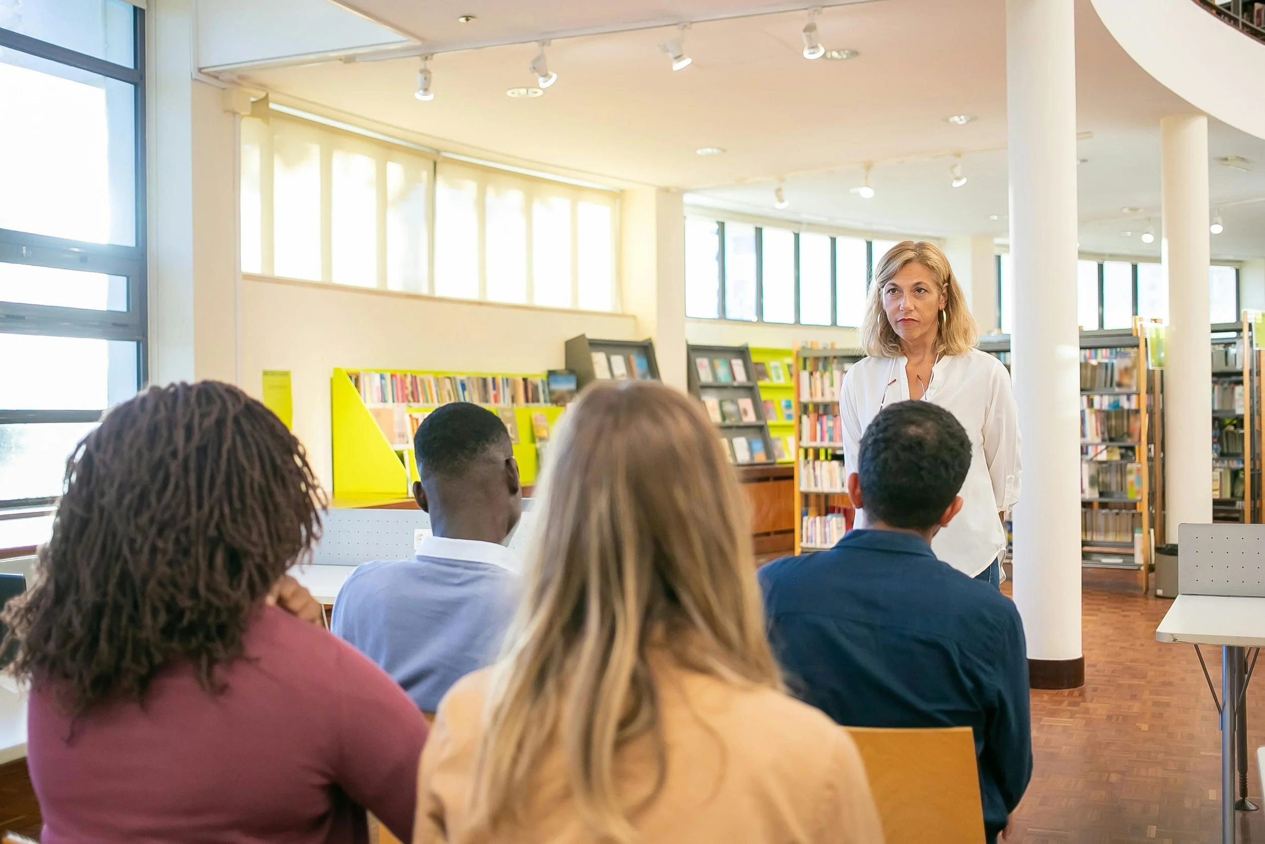 A woman standing in front of a diverse group of four people in a library, presenting a group class