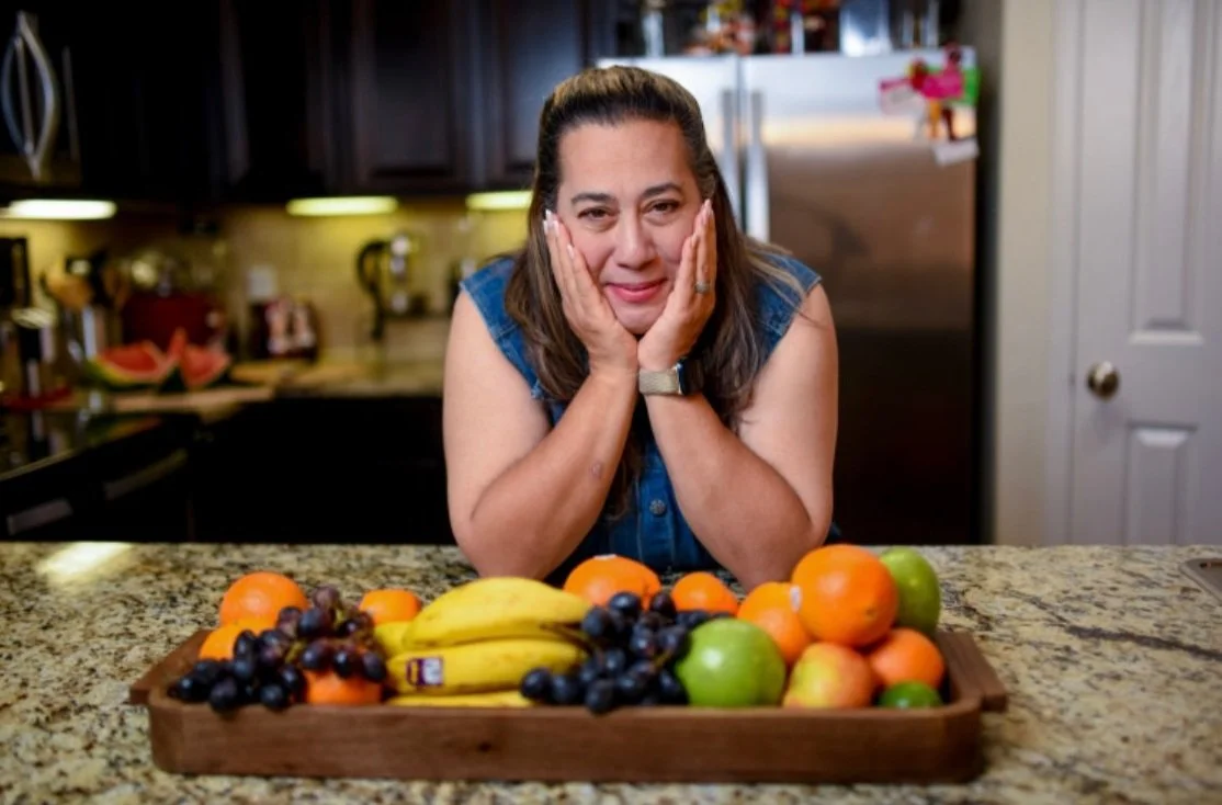Woman with long brown hair, wearing a denim sleeveless top, smiling with hands on her cheeks, sitting at a kitchen counter with a tray of bananas, oranges, apples, and grapes.