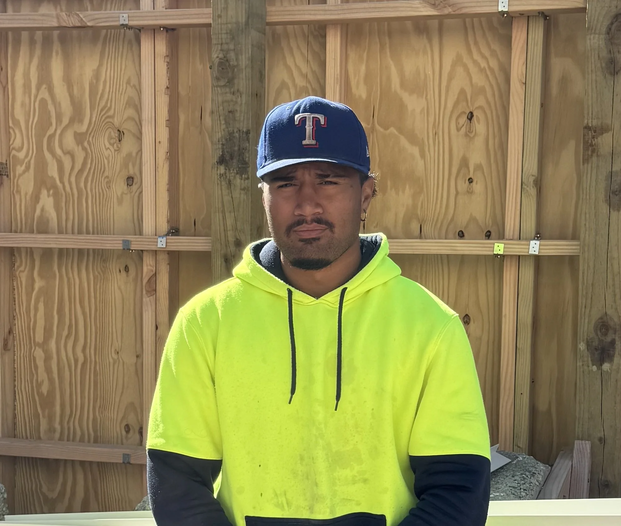 A woman with dark hair styled in a braid, wearing a beige PUMA hoodie, an orange backpack, and holding a magazine or tablet, standing indoors with a metallic and blue background.