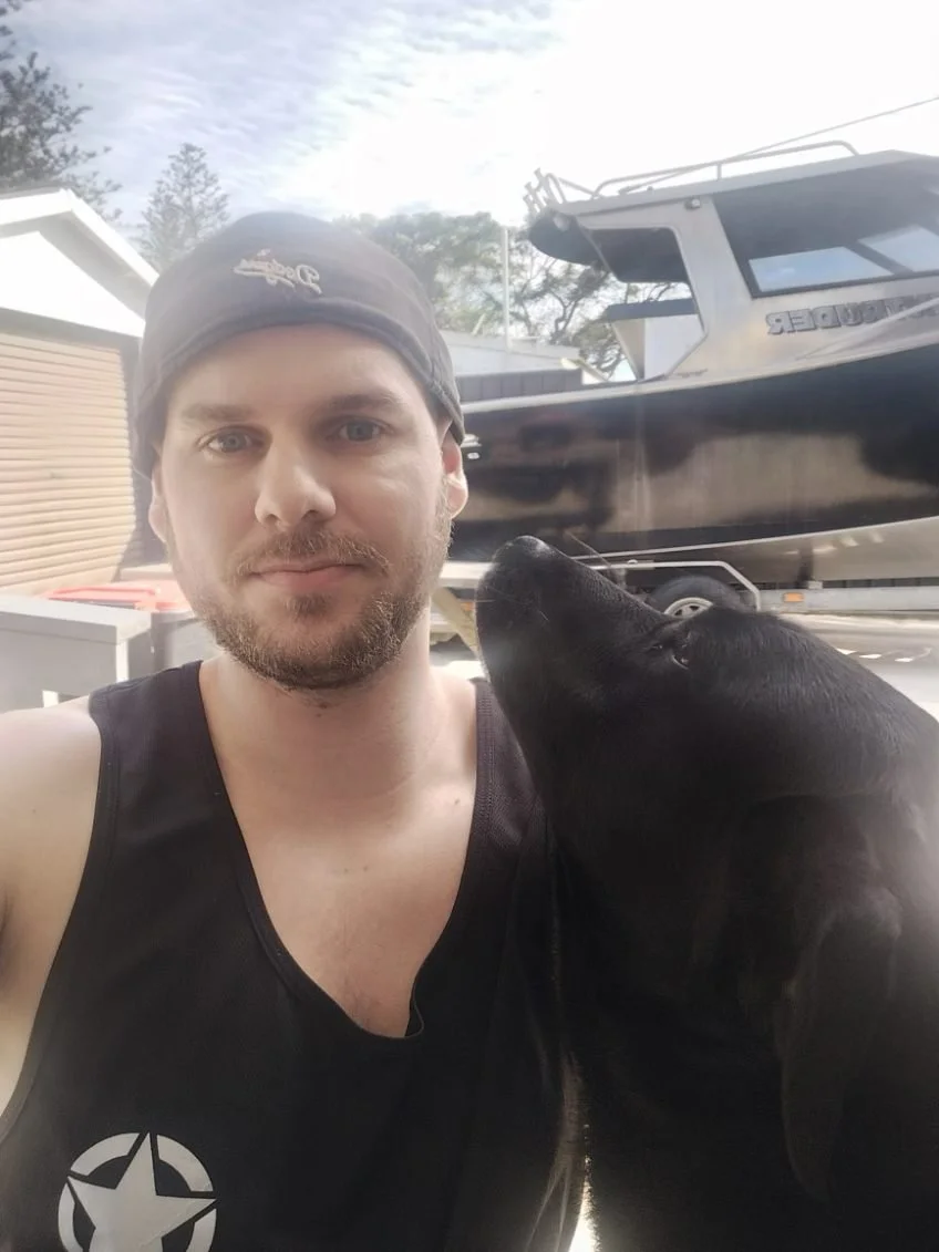 A man giving a thumbs-up in a workshop or lab with electrical equipment, wires, and tools on a table and a green cabinet with tools in the background.